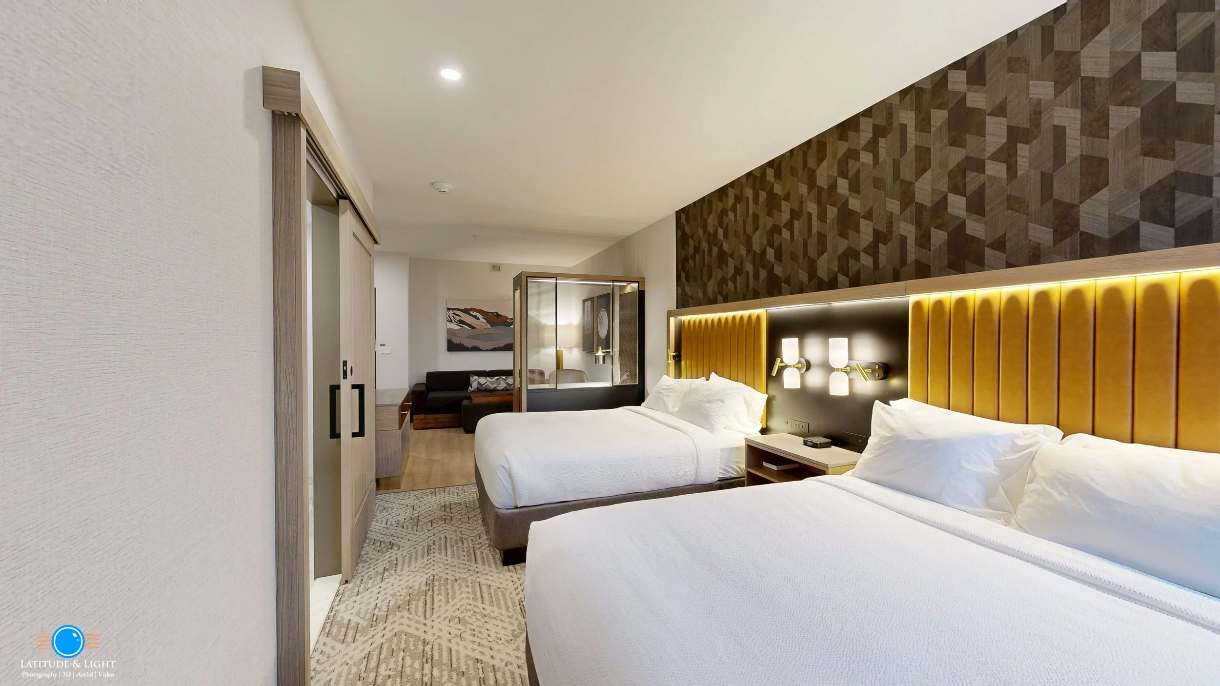 A modern hotel room with two beds with white linens and yellow upholstered headboards, a bedside table with a phone, a wall-mounted light fixture, a patterned carpet, a sliding door, a sitting area with a sofa, a painting on the wall, and a wooden pa