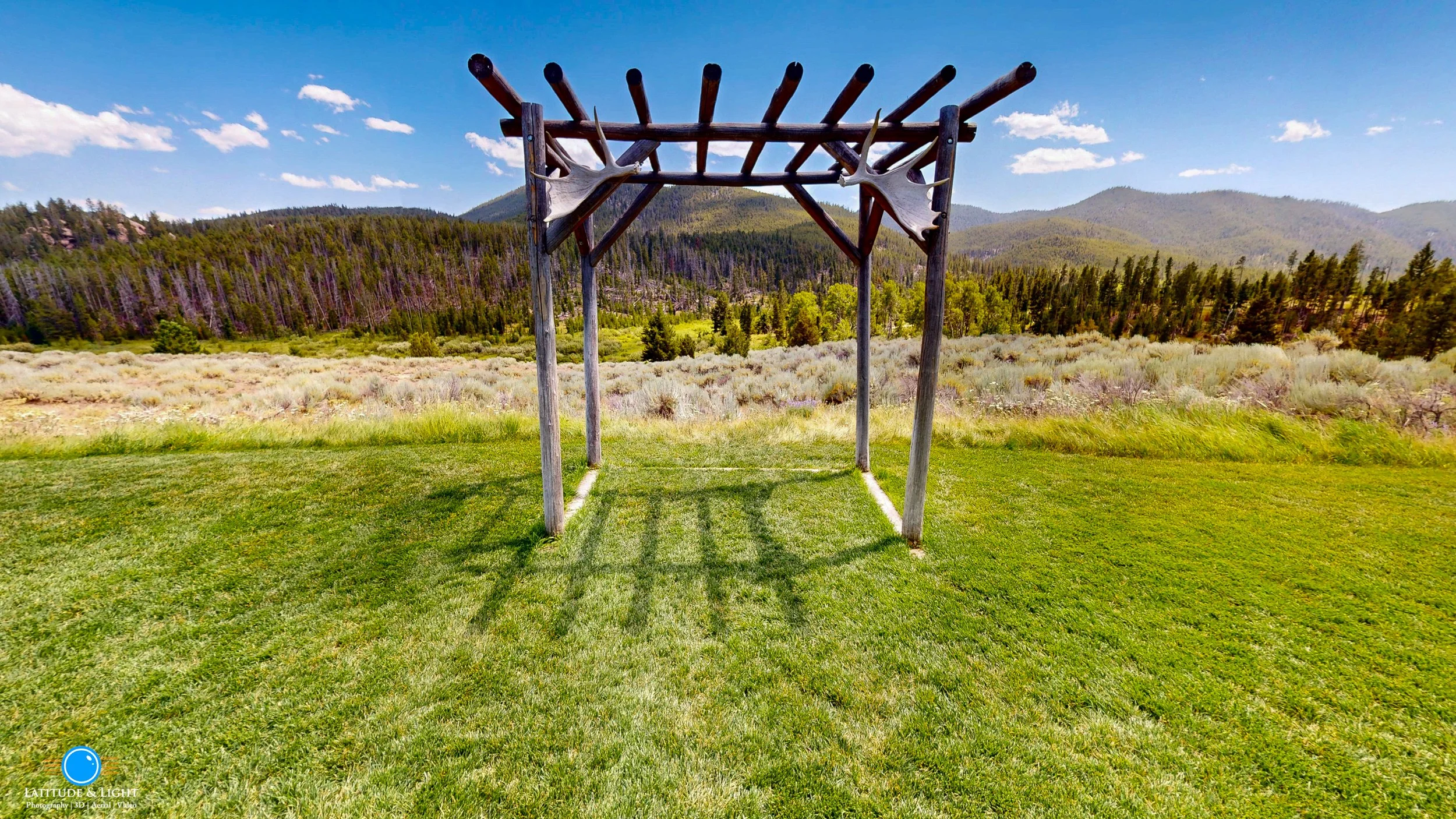 A wooden outdoor ceremony structure with antler decorations on the top beams at a Montana wedding ranch, situated on a grassy lawn with a scenic mountain landscape and forest in the background.