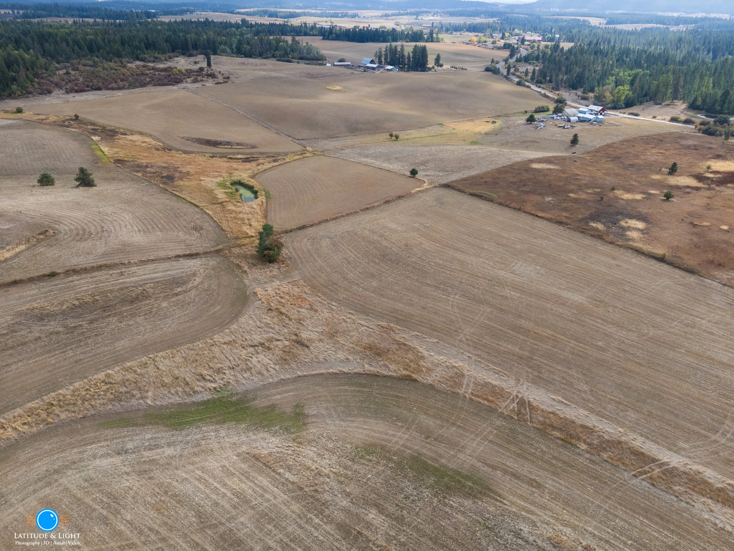 Aerial view of farmland with dry fields, small pond, trees, and farm buildings in the distance.