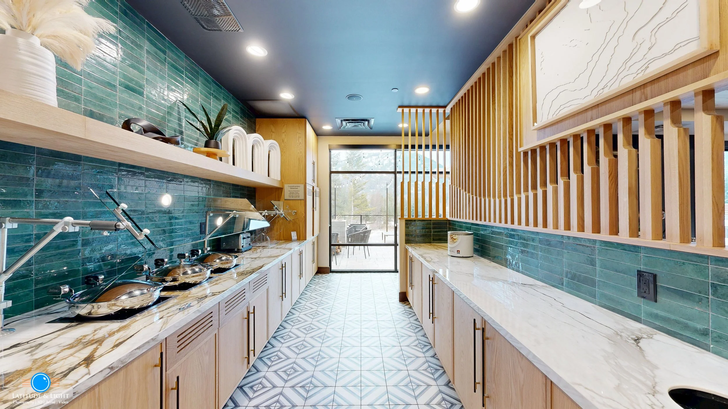  LaQuinta Inn by Wyndham in Walla Walla features a modern buffet area with blue tiled backsplash, marble countertops, light wood cabinets, and a patterned tile floor. Seating area visible through a glass door at the end.