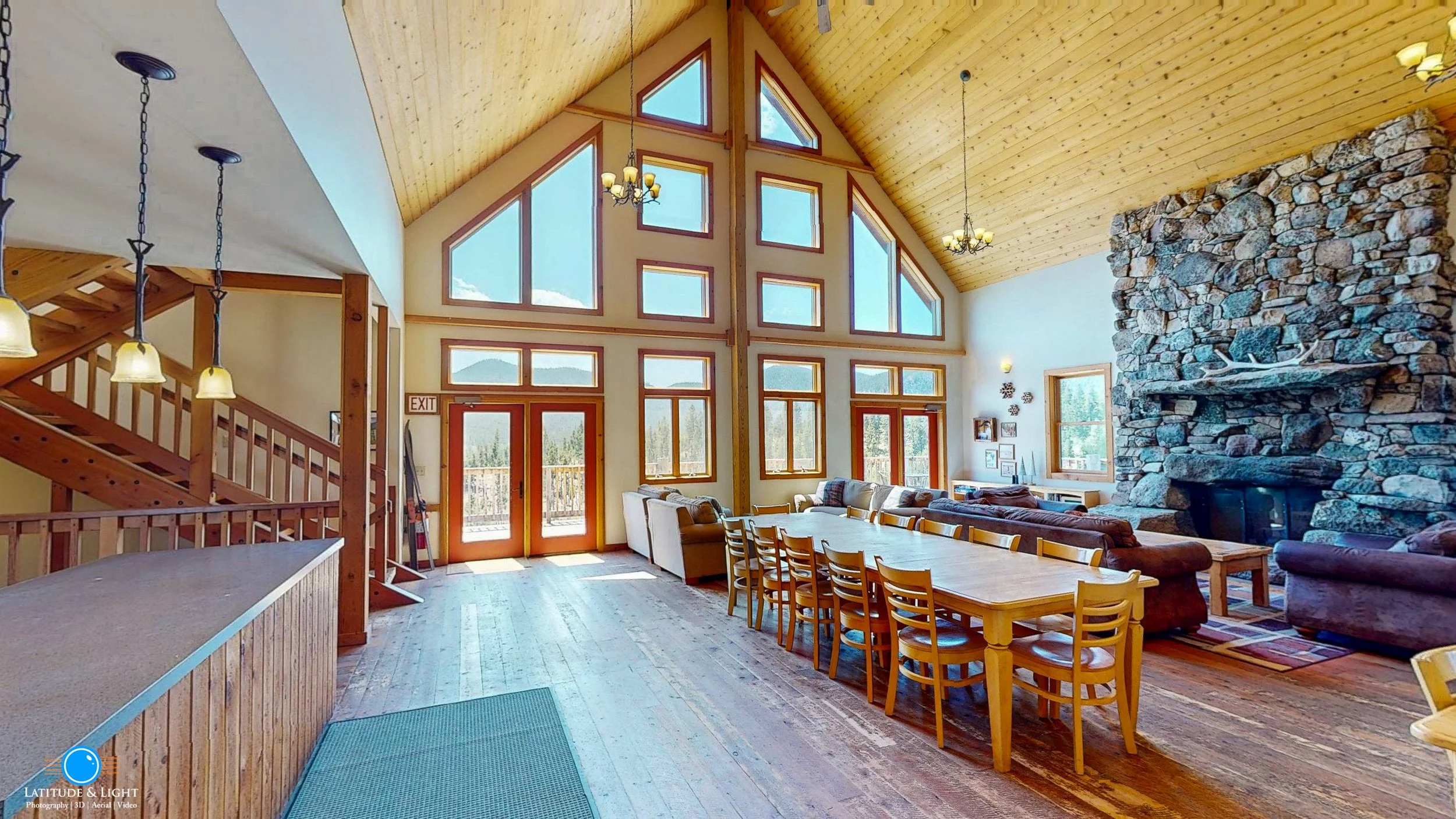 Large living room in a cabin with exposed wooden ceiling, expansive windows, a massive stone fireplace, and a long wooden dining table with chairs.