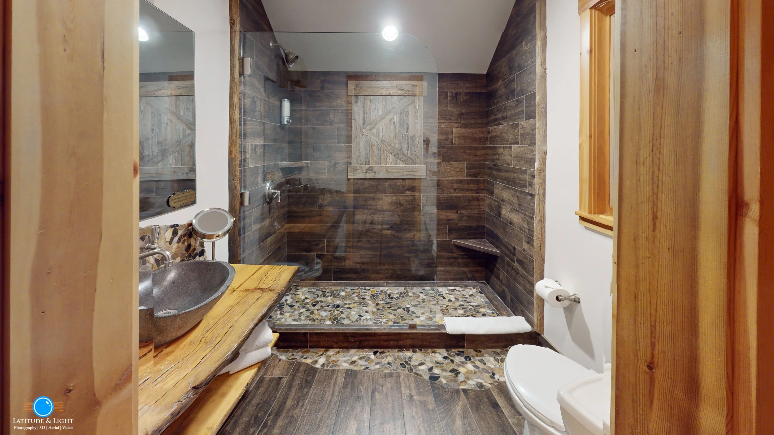 Lake Tahoe bathroom with rustic wood and stone decor, glass shower enclosure with pebble floor, wooden vanity with a gray stone vessel sink, and a toilet with a wooden window on the right side.