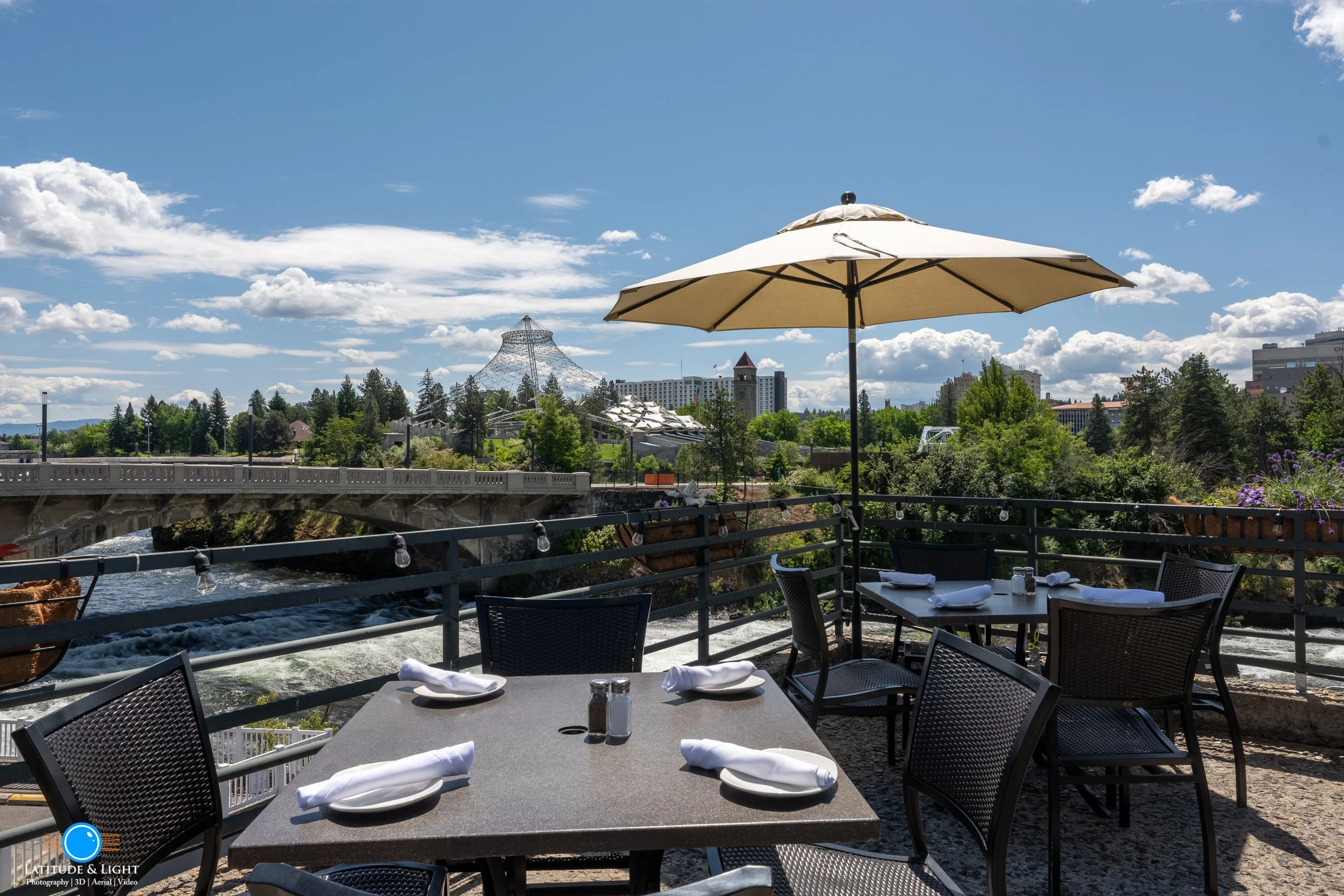 Spokane: Outdoor restaurant patio with tables and chairs under a large umbrella, overlooking a river, with trees and city buildings in the background, under a partly cloudy sky.