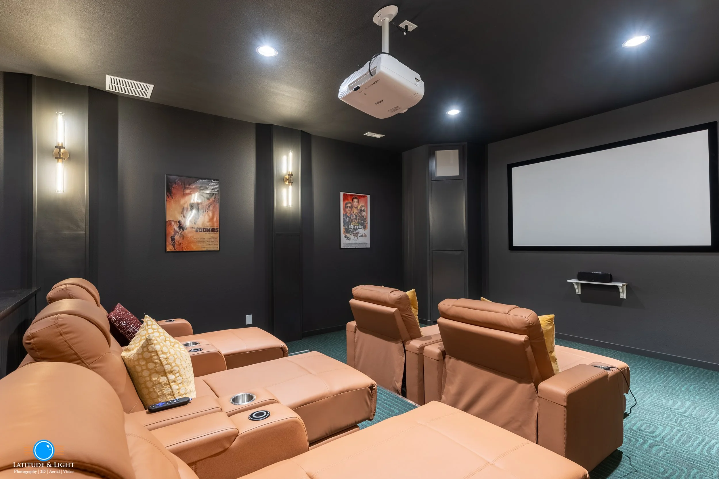 At an apartment complex near Spokane, a home theater room with a large blank white projection screen on a dark wall, beige reclining theater seats, movie posters on the walls, and a ceiling-mounted projector.