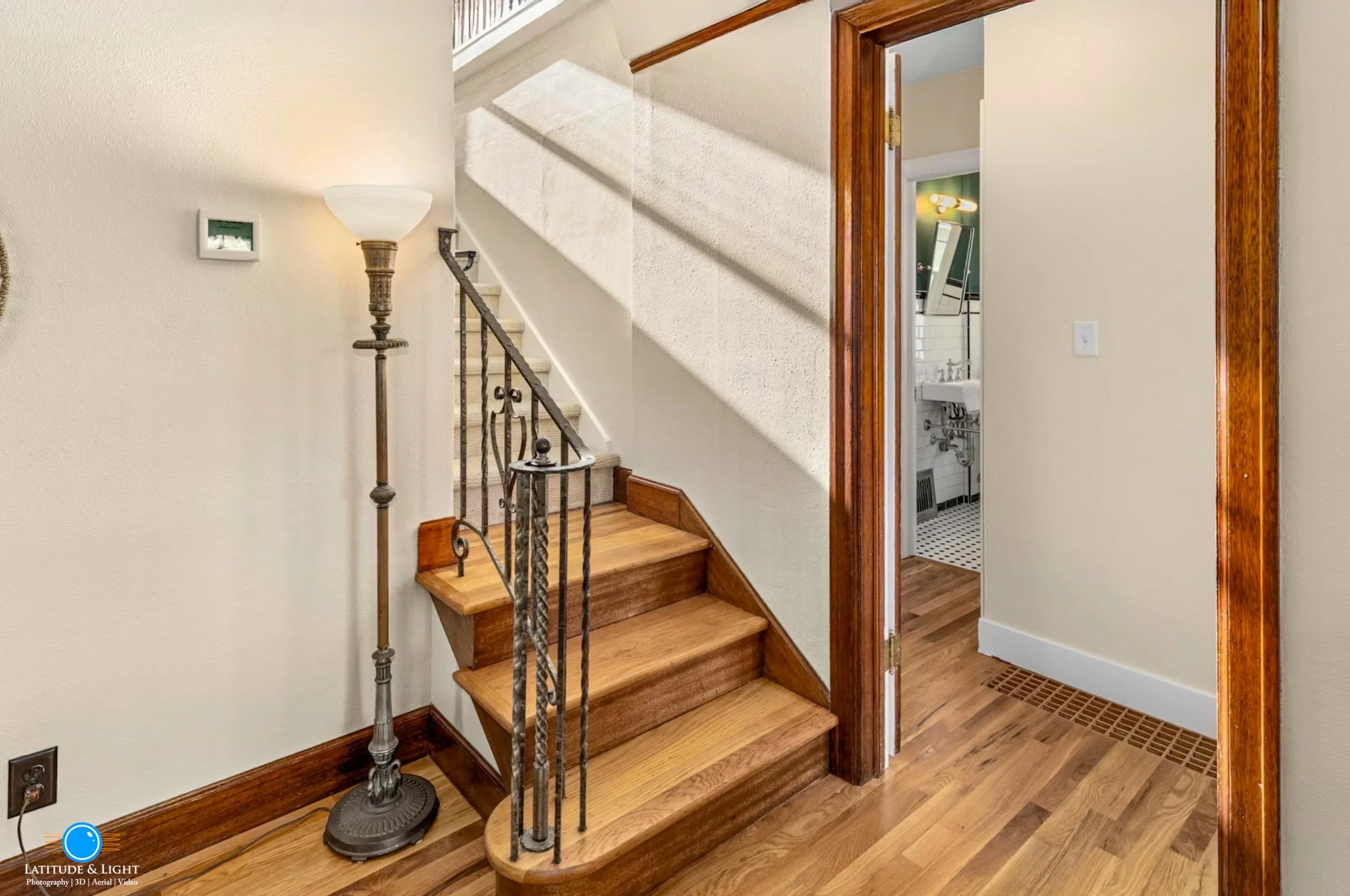 Interior view of a house showing a staircase with a metal railing, a wooden floor, a wall-mounted thermostat, a tall floor lamp, and a doorway leading to a bathroom with a sink and a mirror.