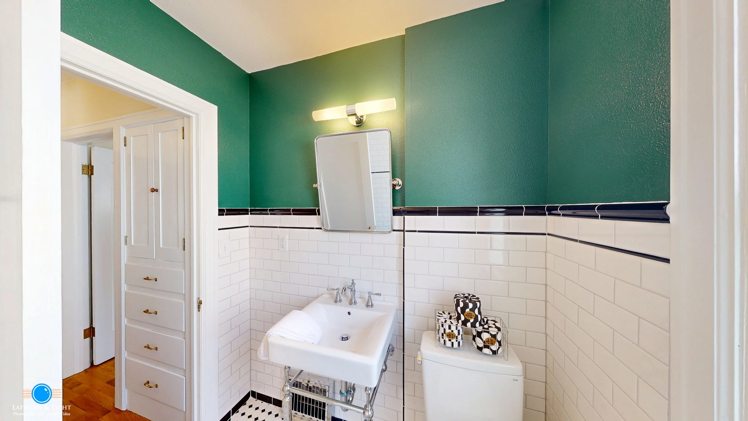 Bathroom with a green upper wall, white tiled lower wall with black trim, small white sink, mirror, and toilet with black and white tissue boxes on top.