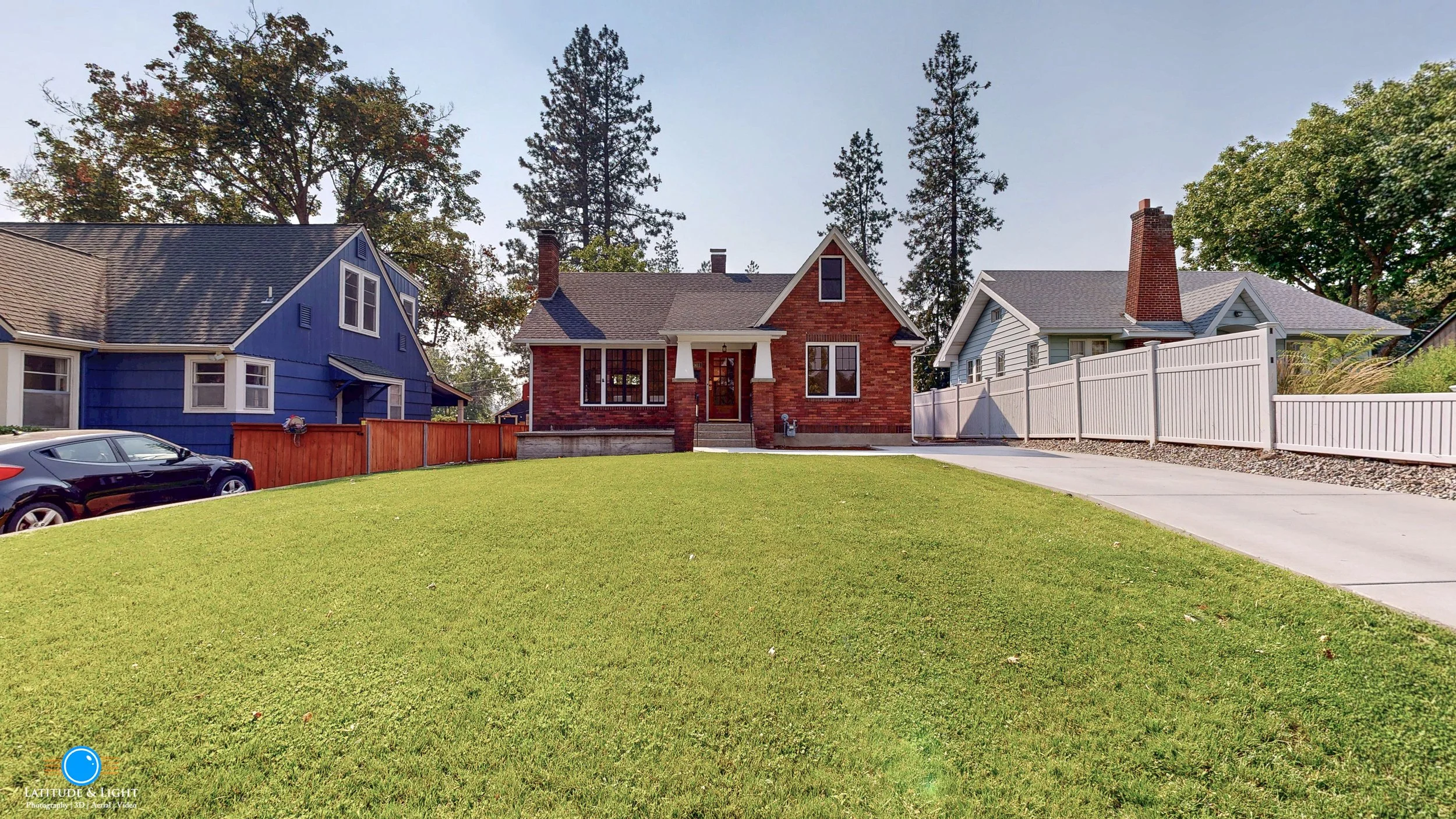 A well-maintained front yard looking towards the home entrance in Spokane. There is a well-maintained green lawn in front and a concrete driveway on the right. The house is surrounded by trees and neighboring houses with different colors and fences.