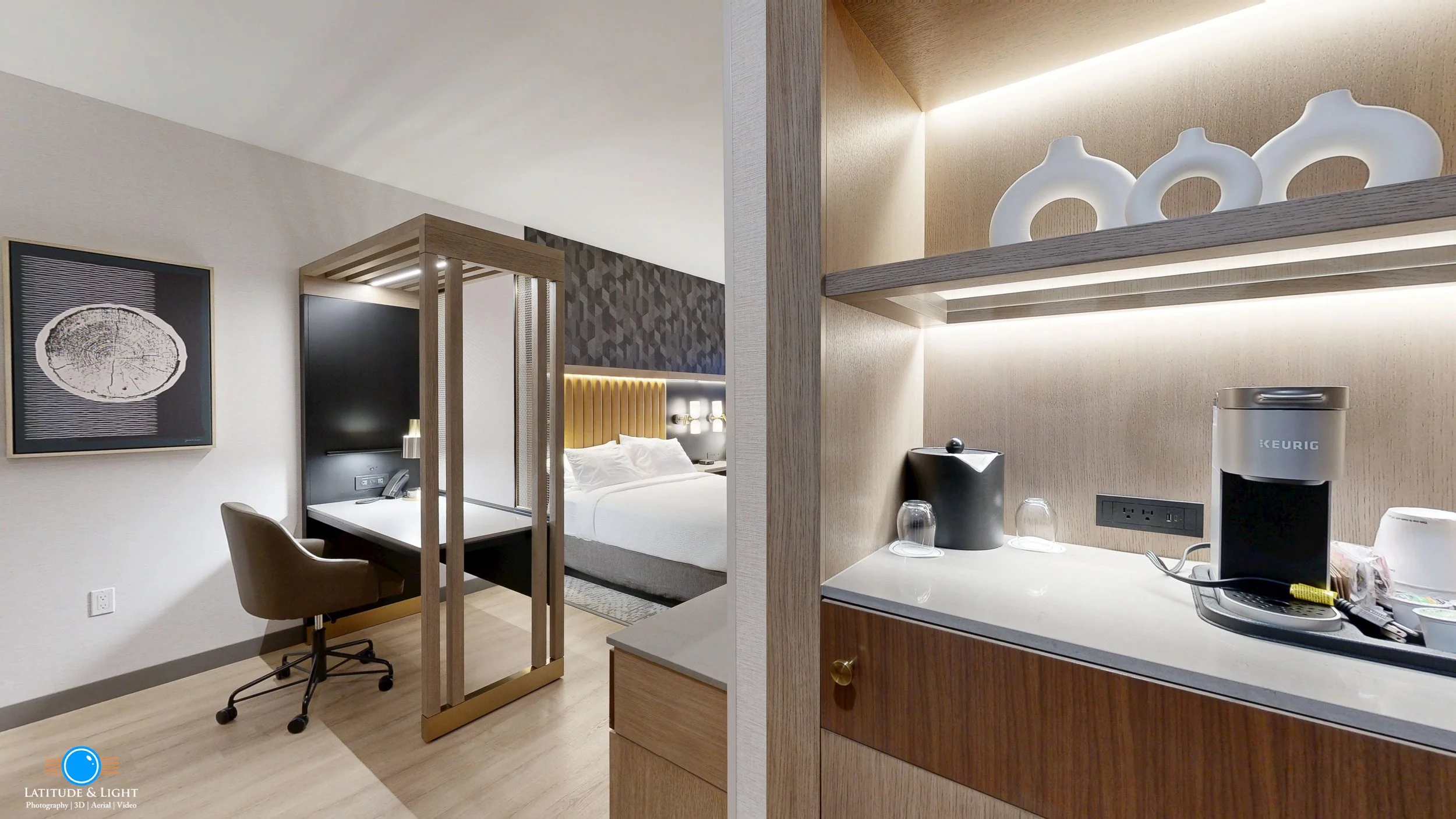 A hotel room with a workspace, a bed with a padded headboard, and a kitchenette area with a Keurig coffee maker and decorative vases on a shelf.