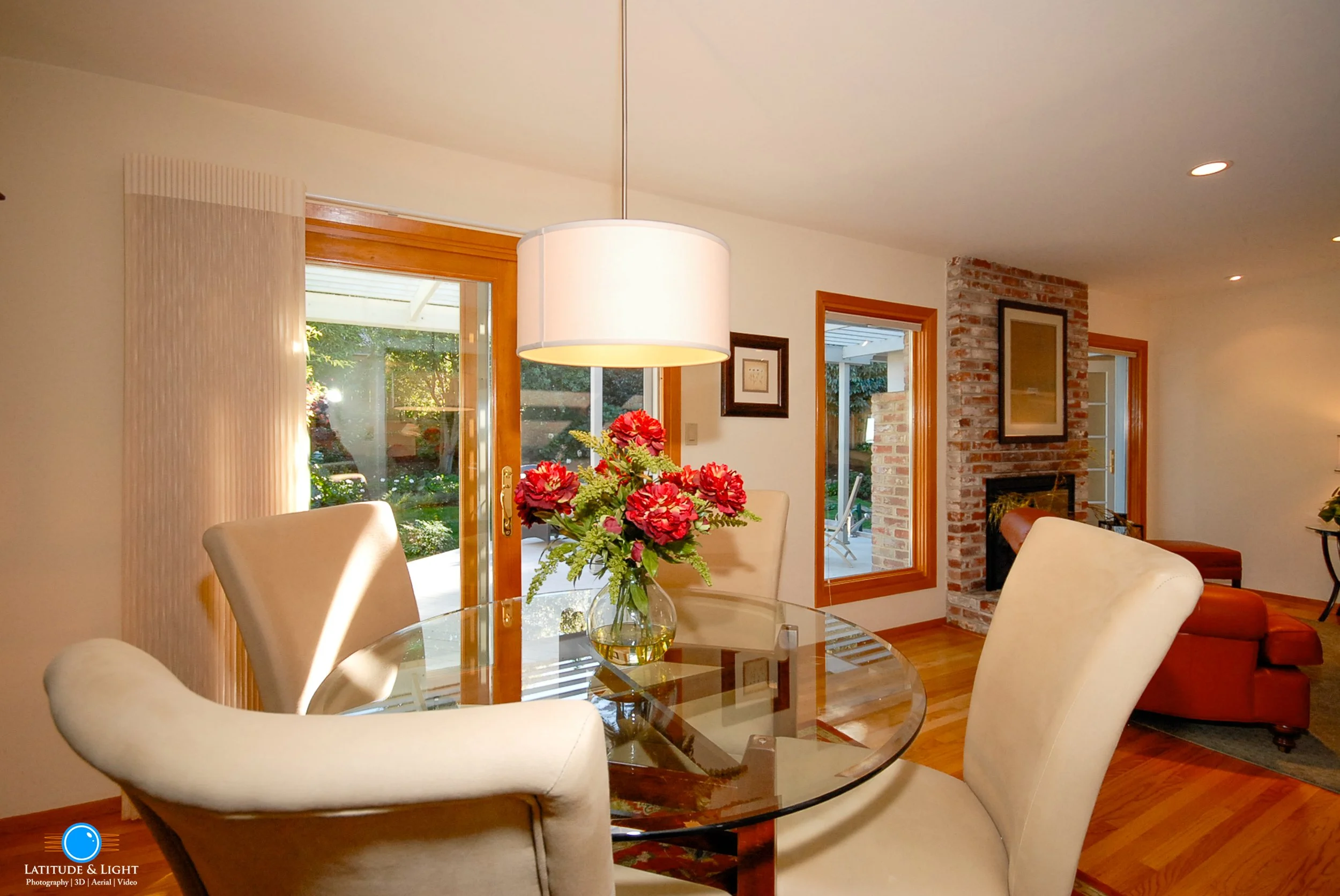 A dining room with a round glass table, cream-colored chairs, a vase with pink flowers, a large white hanging light fixture, sliding glass door, brick fireplace, and a view of a patio and garden outside.