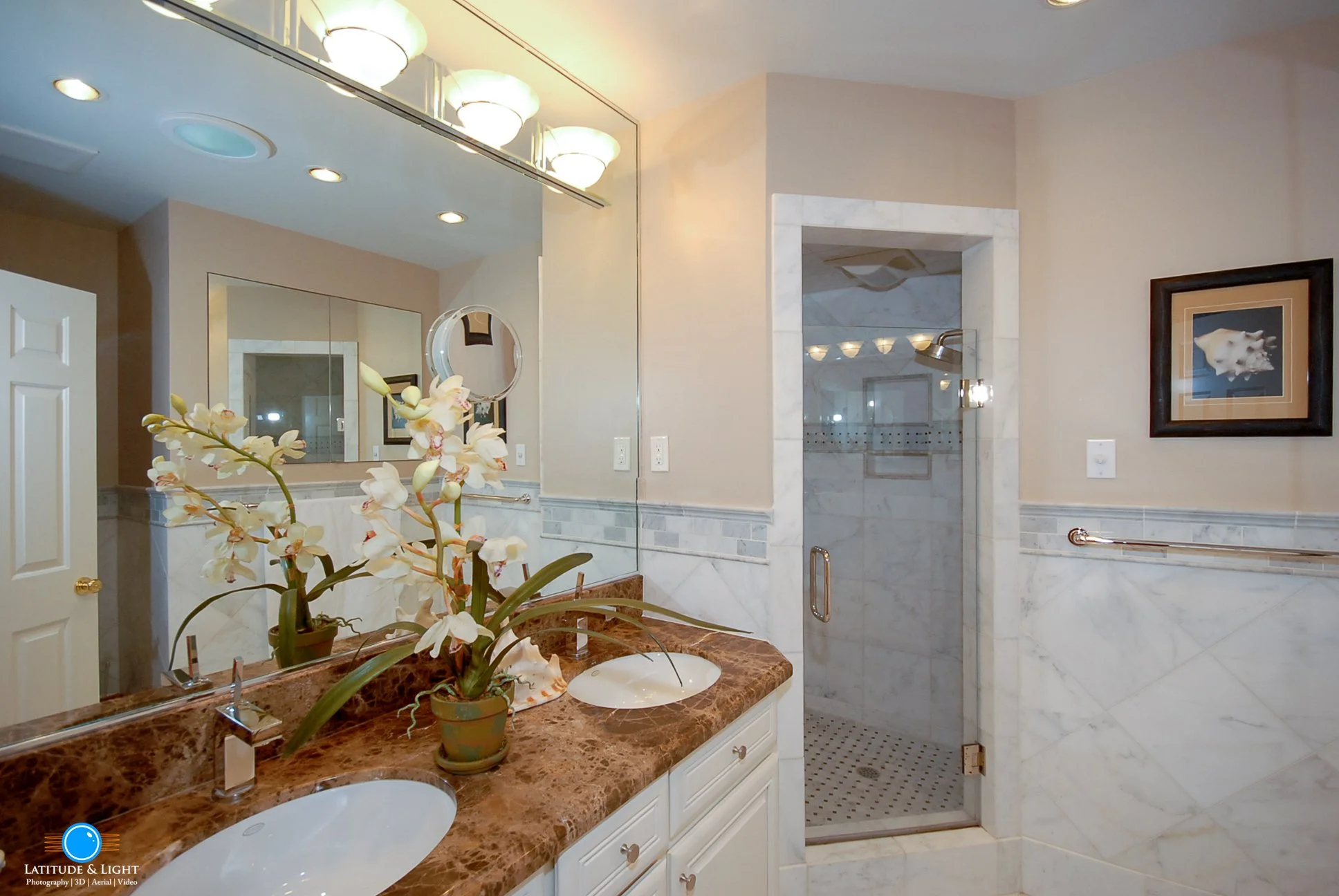 Luxury bathroom in Coeur D'Alene with dual sinks on a marble countertop, a large mirror above, a potted orchid plant, a glass-enclosed shower, framed artwork, and cream-colored walls.