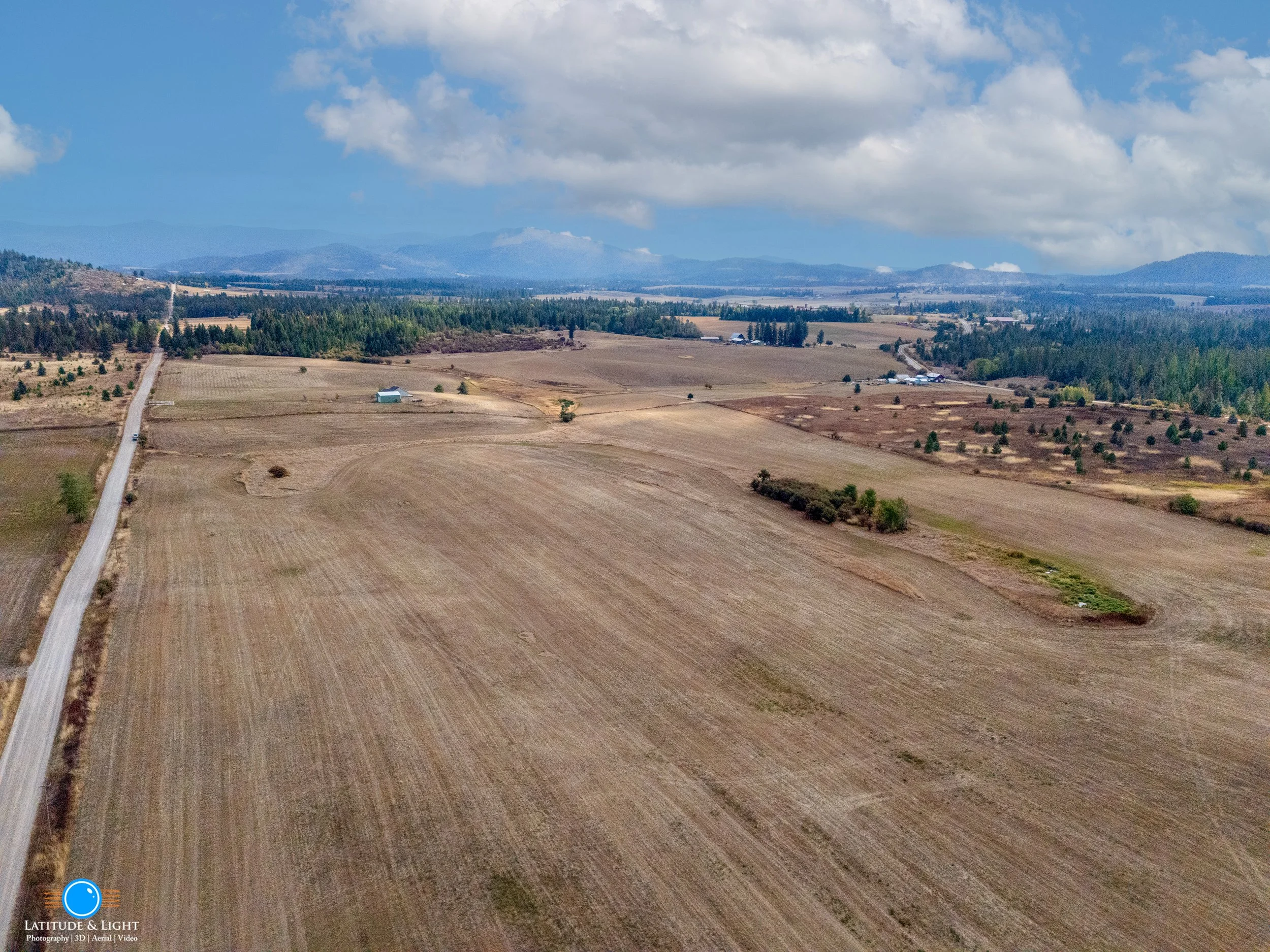 Aerial view of a rural farmland landscape with a narrow road running along the left side, open fields, scattered trees, farm buildings in the distance, and mountains under a cloudy sky.
