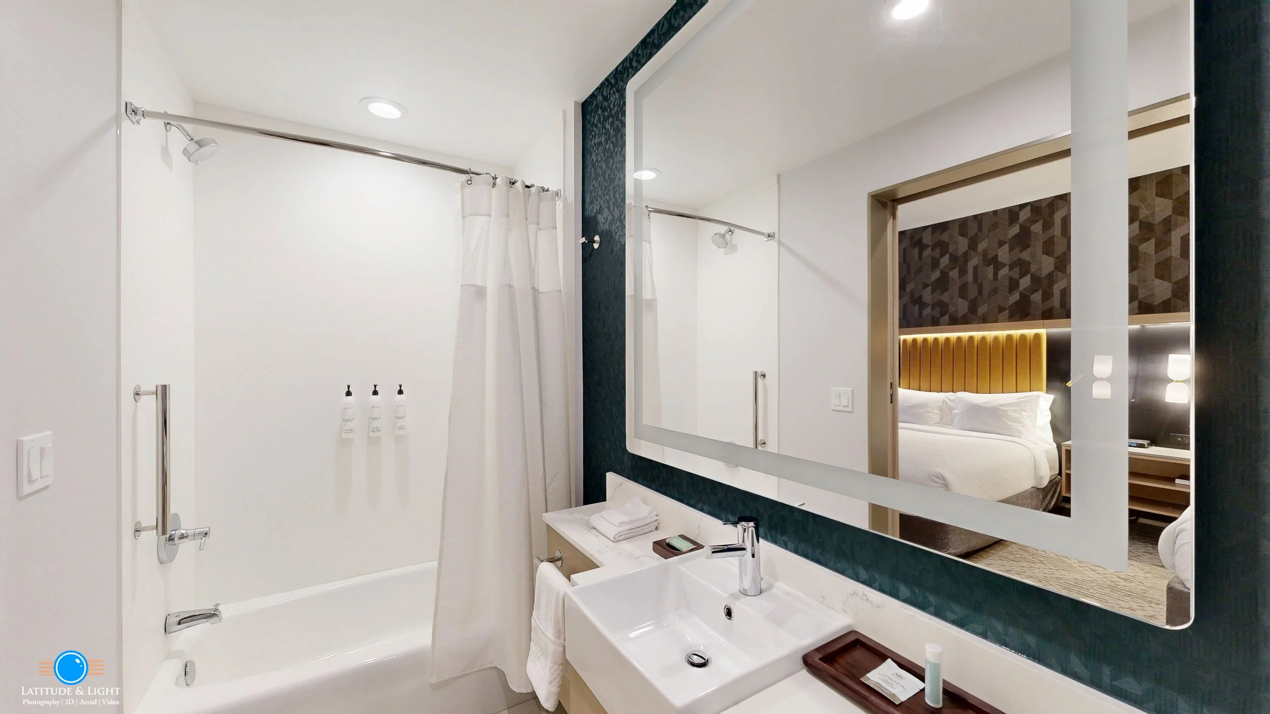 Modern hotel bathroom with a shower area, white shower curtain, large mirror, white sink, and view of a bedroom with a bed and decorative headboard