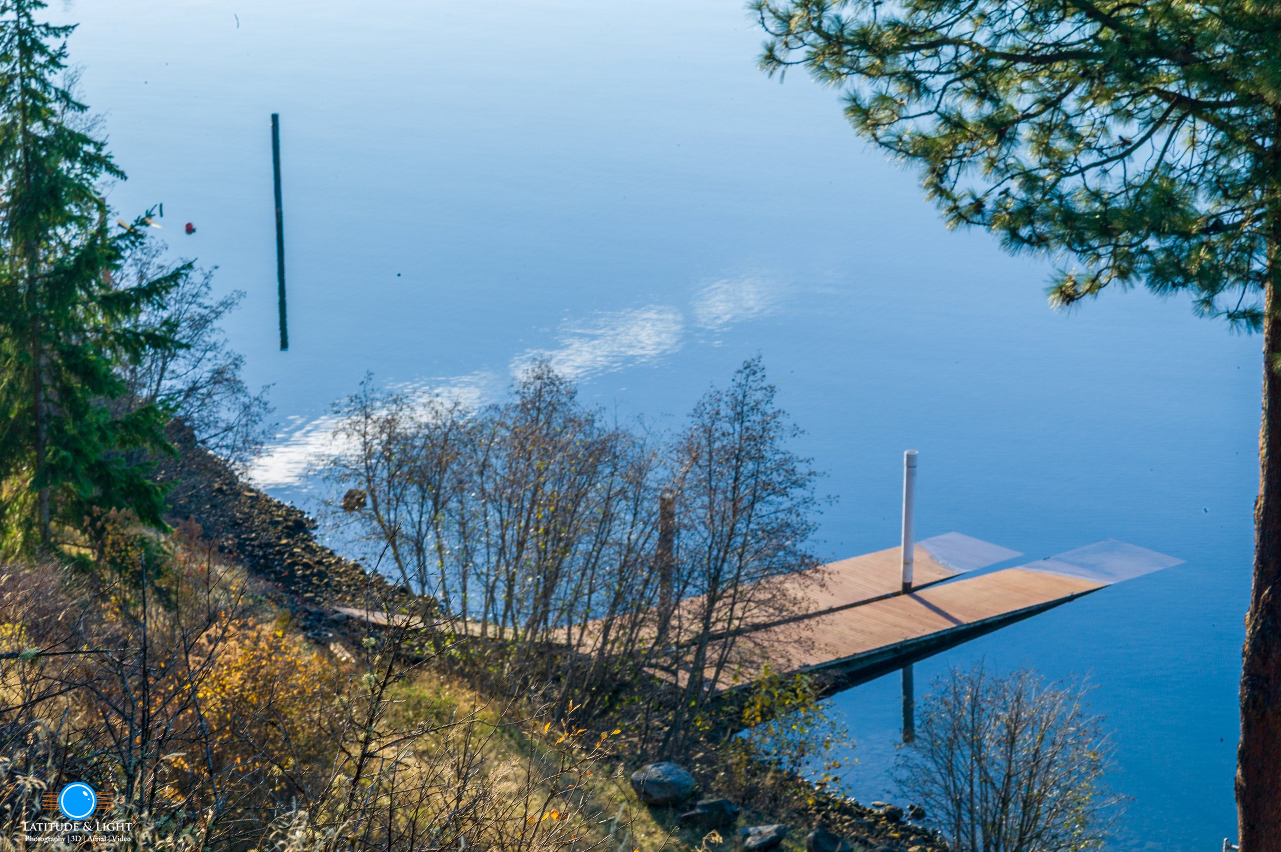 Harrison and Lake Coeur D'Alene: A tranquil lakeside scene with calm water reflecting the sky and surrounding trees, a partially submerged wooden dock with a white pole, and a mix of evergreen and leafless trees along the shore.