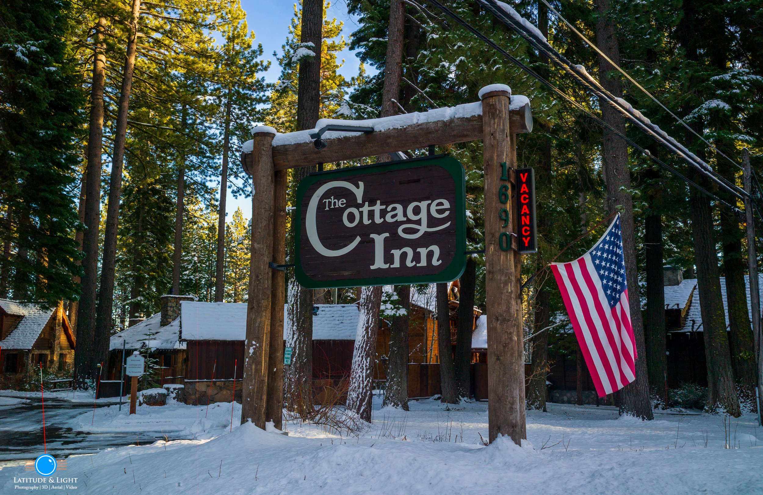 A wooden sign for The Cottage Inn at Tahoe City, with a neon sign showing 'VACANCY' in red letters, and an American flag hanging beside it. The background features snow-covered ground, tall trees, and cabins in a forested area.
