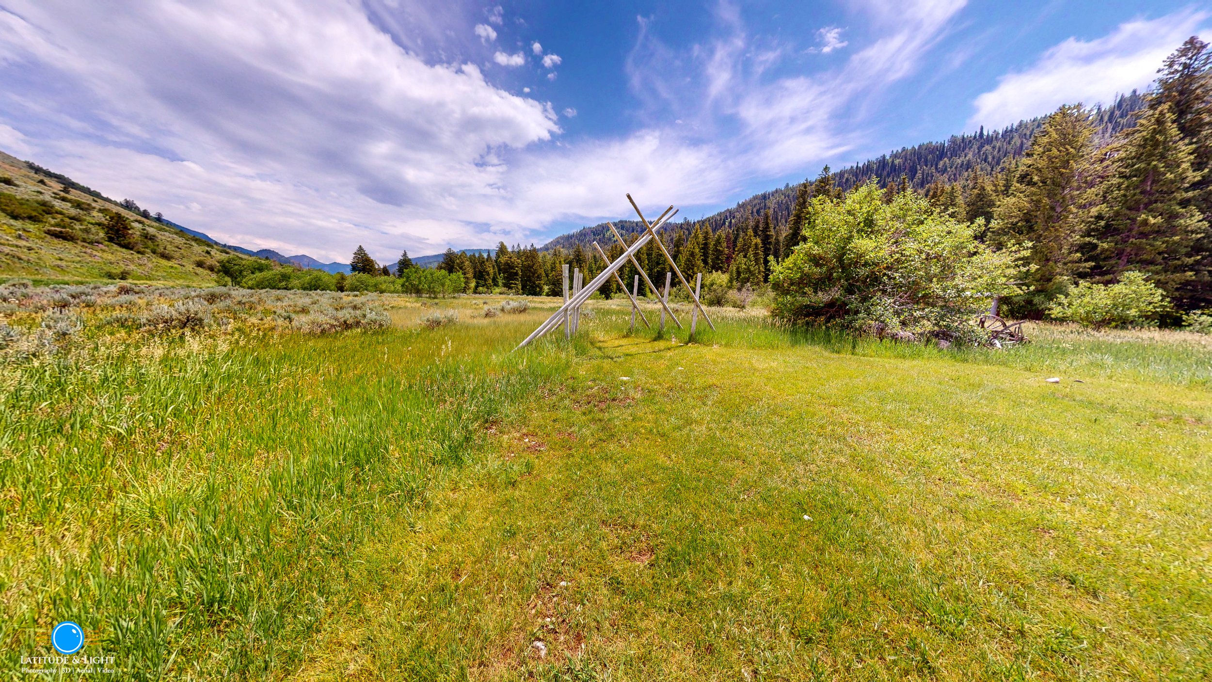 A ceremony canopy at a Montana ranch sits in an open grassy field near a green bush, surrounded by trees and mountains under a partly cloudy sky.
