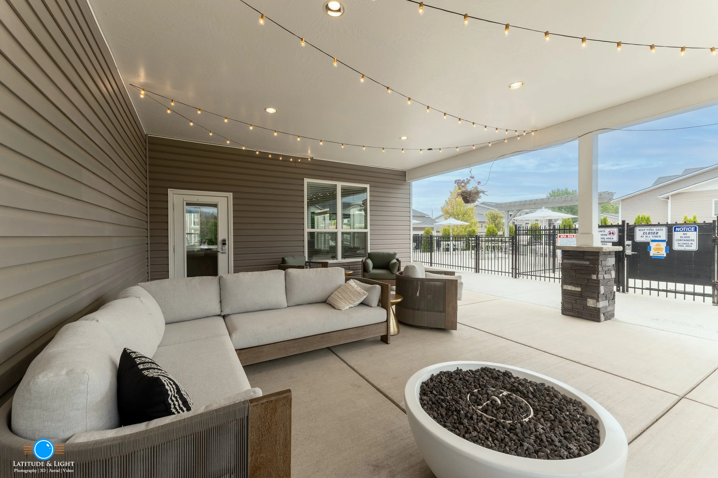 Covered patio with string lights at a Spokane Valley community center has a large beige sofa with black and beige pillows, a set of seating chairs, a white fire pit, and a view of a fenced outdoor pool area with umbrellas and lounge chairs.