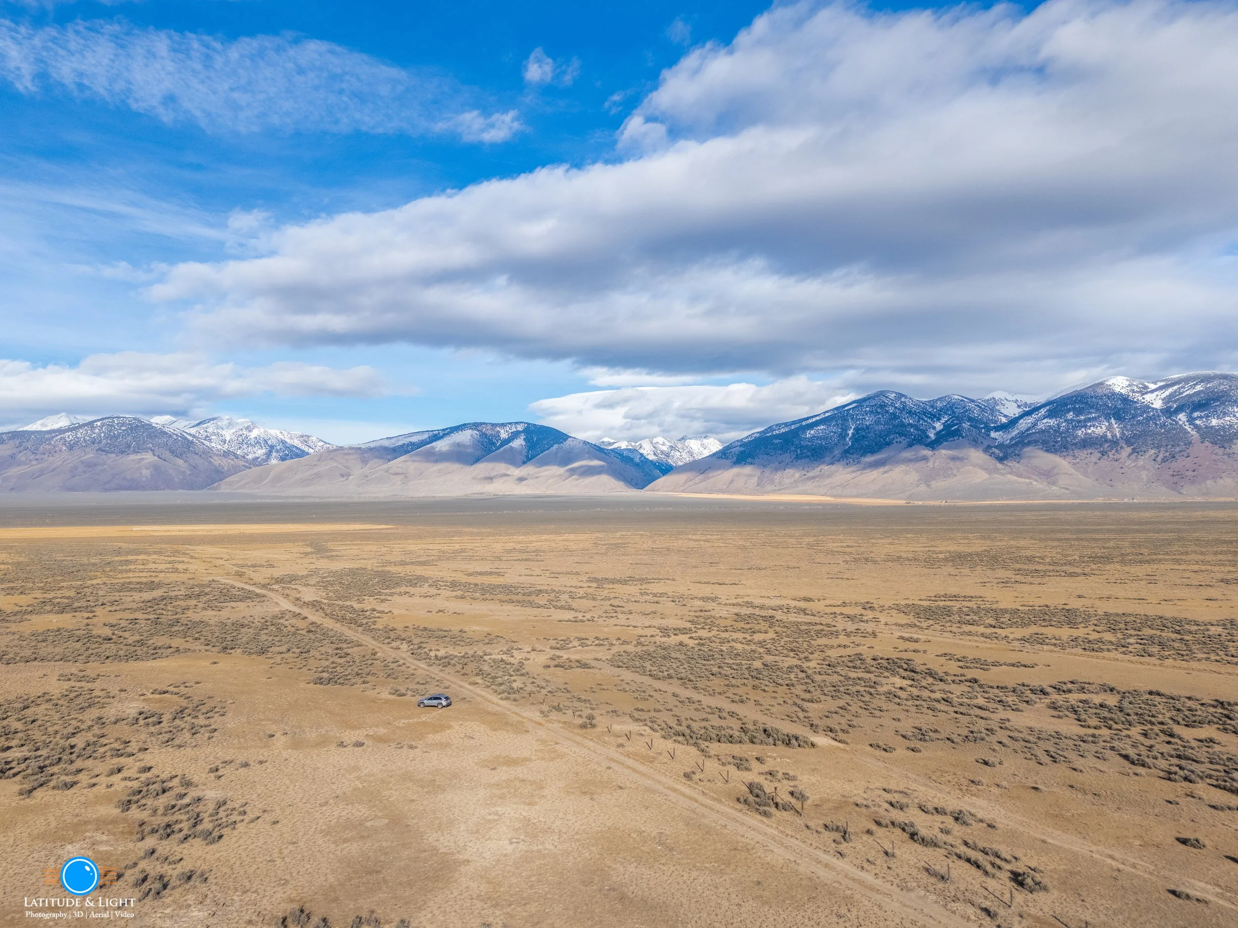 A vast desert landscape with dry, sparse vegetation, a single vehicle on a dirt road, and mountains with snow-capped peaks in the background under a partly cloudy sky.