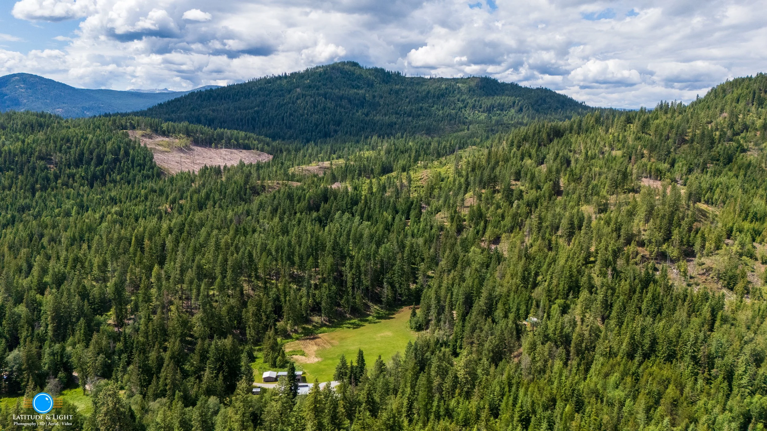 Harrison, Idaho: An aerial view of a lush green forested mountain landscape with a small farm at the bottom, tents, and a large open grassy area. The sky is partly cloudy.