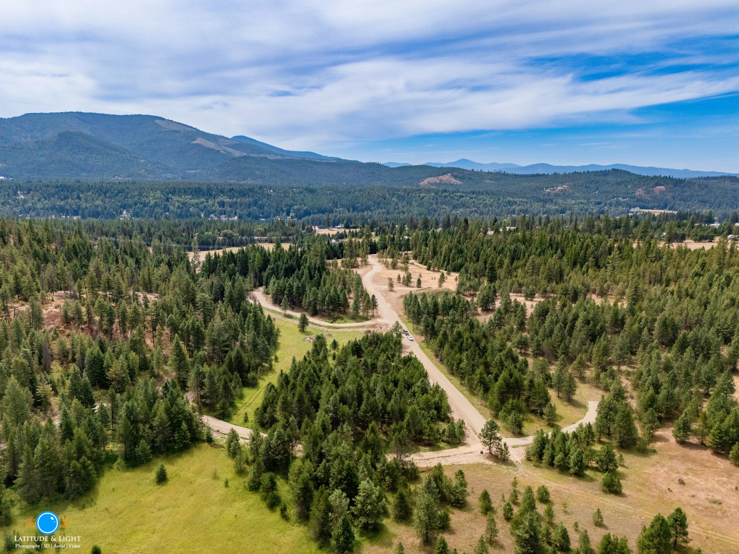 Aerial view of a forested landscape with dirt roads, surrounded by mountains and a partly cloudy sky.