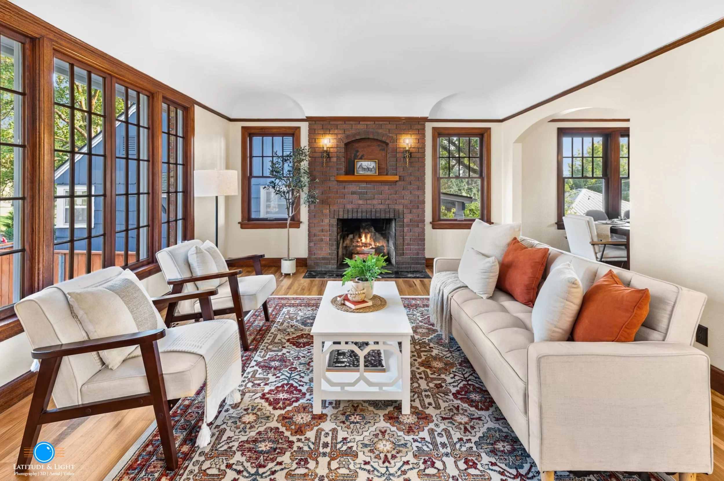 Living room in a pre-war Spokan home with beige sofa, two beige armchairs with wooden arms, a white coffee table, a patterned rug, a brick fireplace, and large windows with wooden trim.