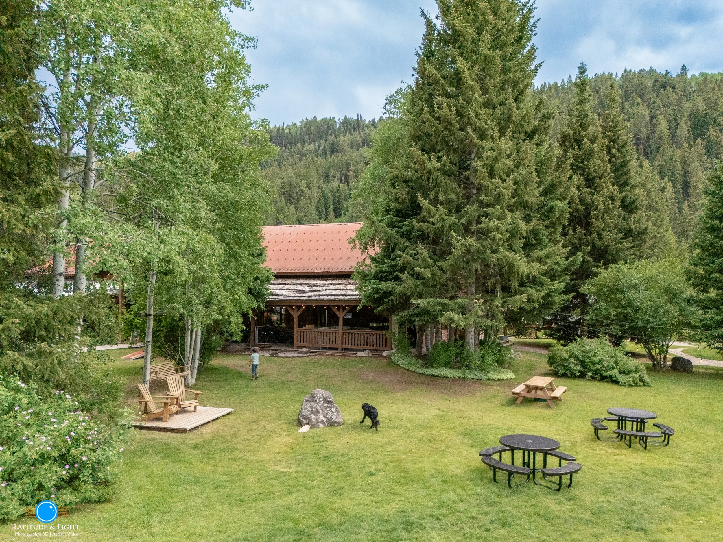 North Idaho land for sale includes a grassy outdoor area with trees, wooden benches, picnic tables, and a building with a red roof in the background, hillside with trees, and a person walking with a dog.