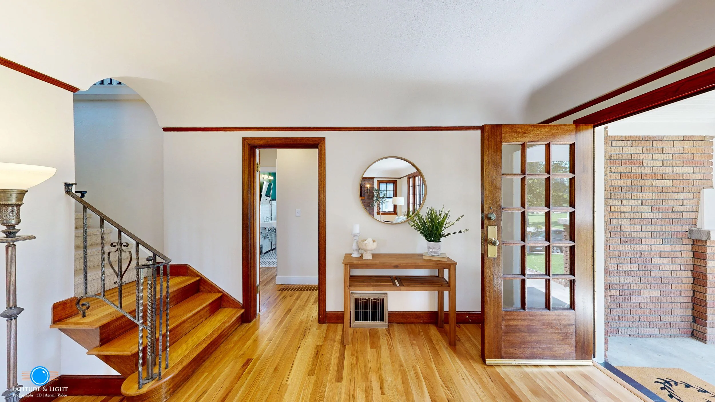 Entryway with wooden floors, wooden door with glass panes, stairs with wrought iron railing, small console table with decorative items, round mirror on wall, window showing outside scene with grass and trees.