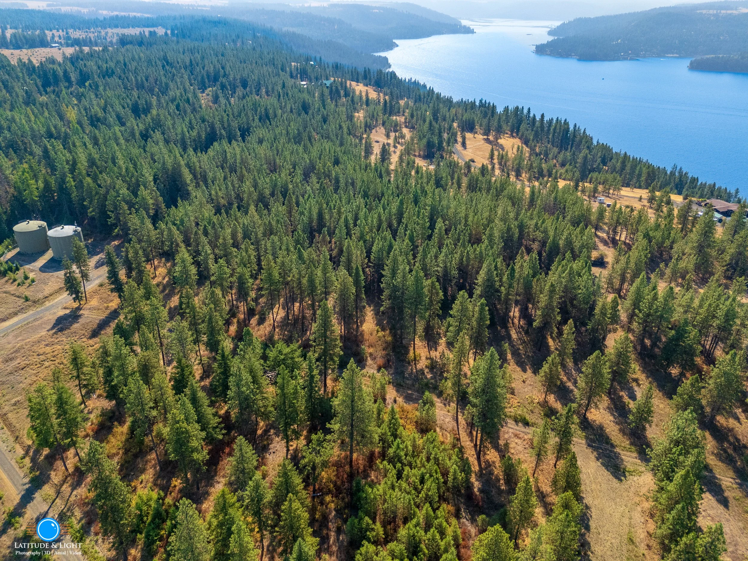 Land in Harrison, Idaho: Aerial view of a forested area near a large body of water with trees, water, and dry patches of land, along with some structures and a road.