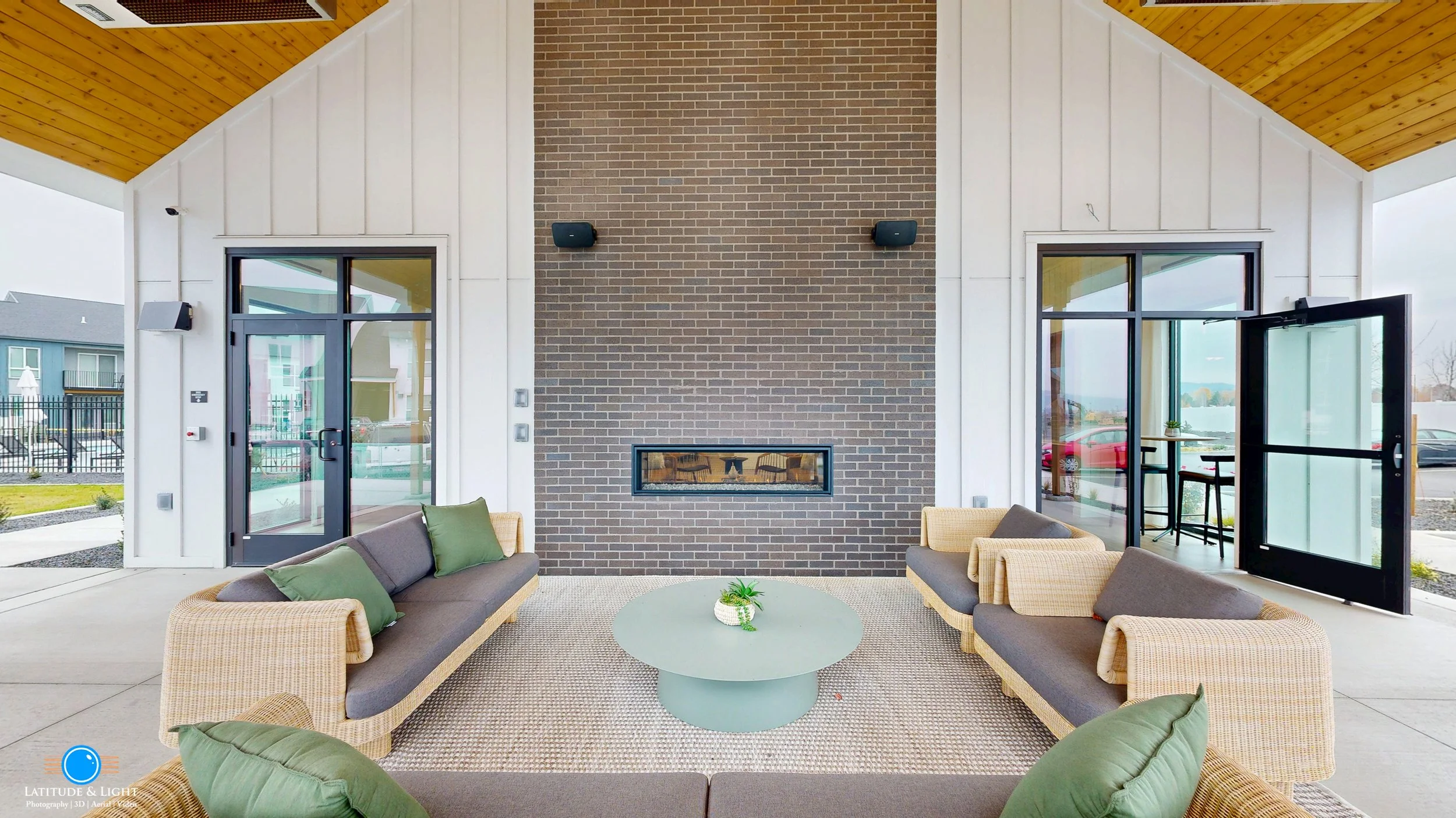 Outdoor lounge area at Spokane Valley area apartment community center  with two sofas, green pillows, a round coffee table, and a brick fireplace on the back wall, with sliding glass doors and windows on either side.