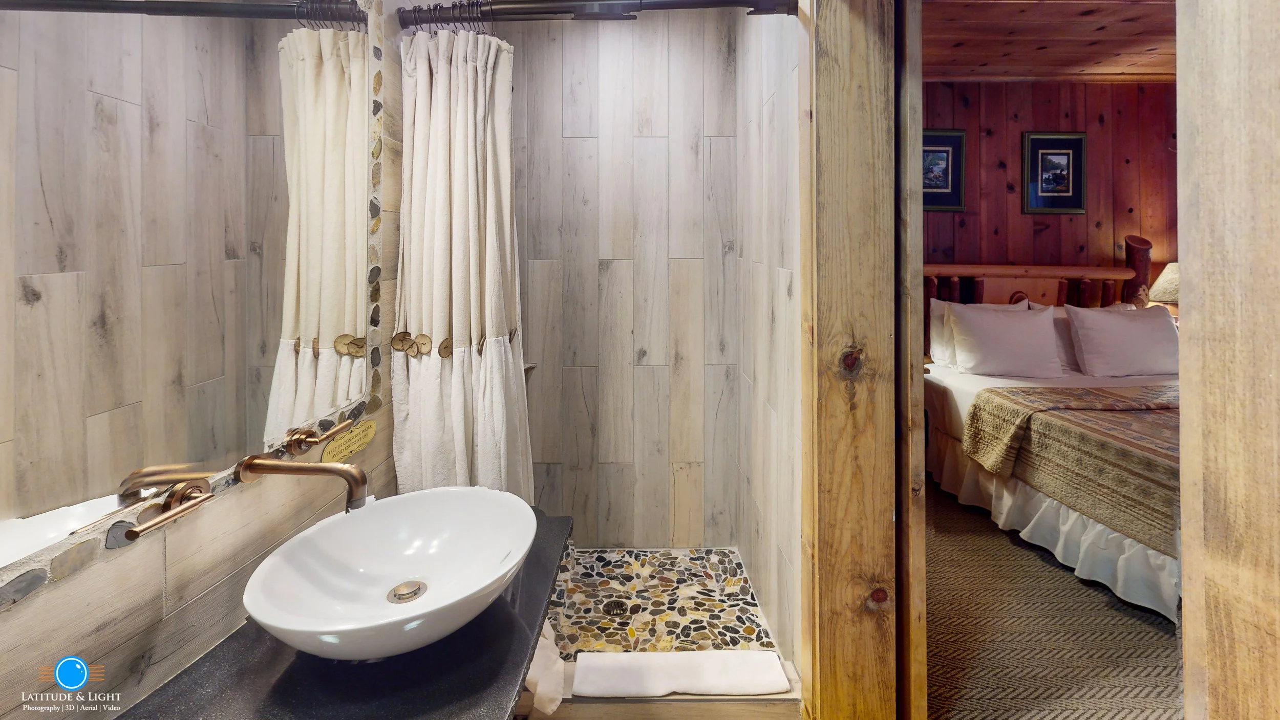Bathroom with stone textured walls, a white sink with a copper faucet, and a shower area with a pebble tile floor, partially obscured by a curtain. Adjacent to the bathroom is a bedroom with a wooden headboard, white pillows, and framed pictures on a