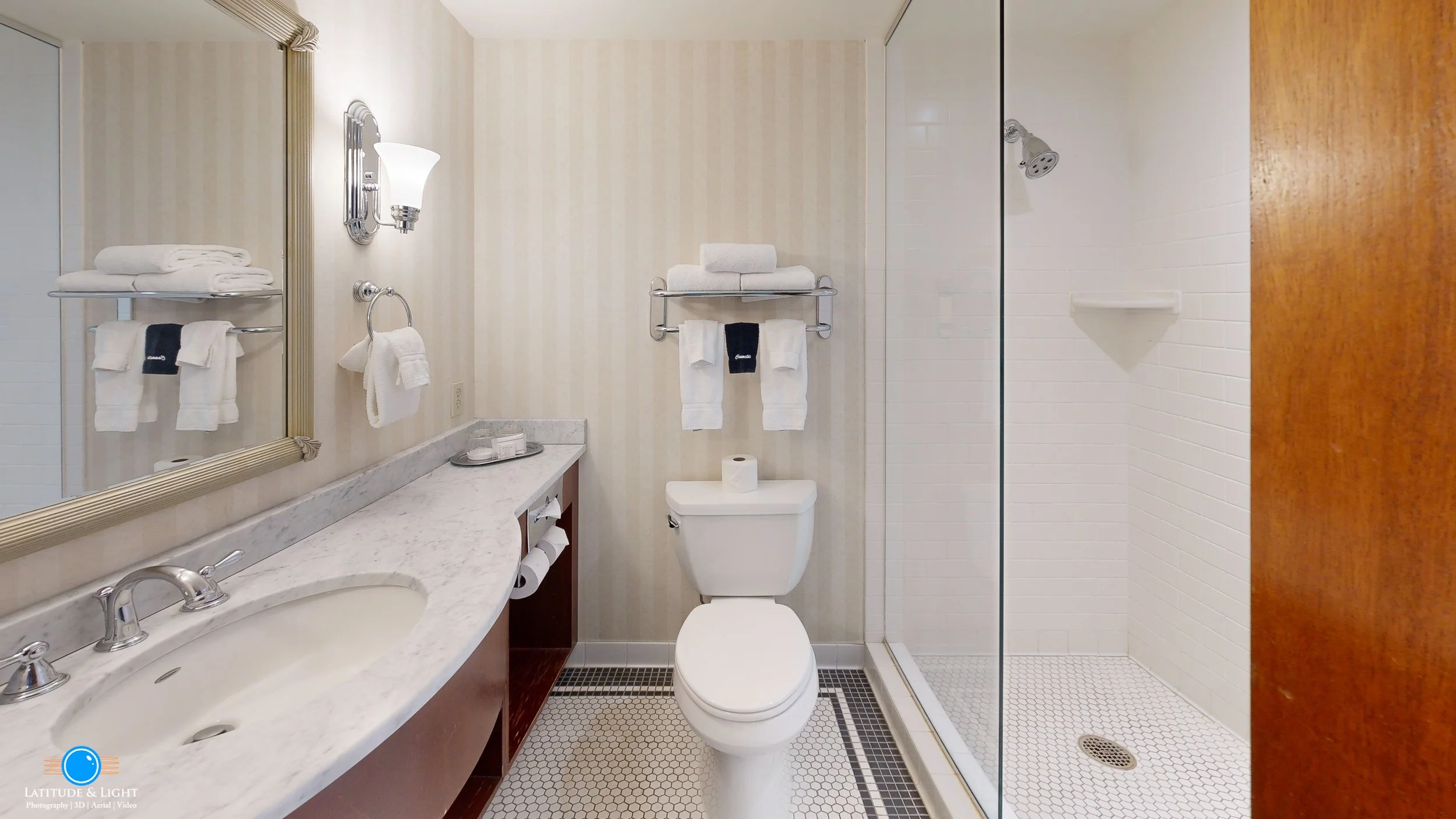 Bathroom with a marble vanity, large mirror, and towel rack, a toilet with a roll of toilet paper, and a walk-in shower with white subway tiles and a small shelf.