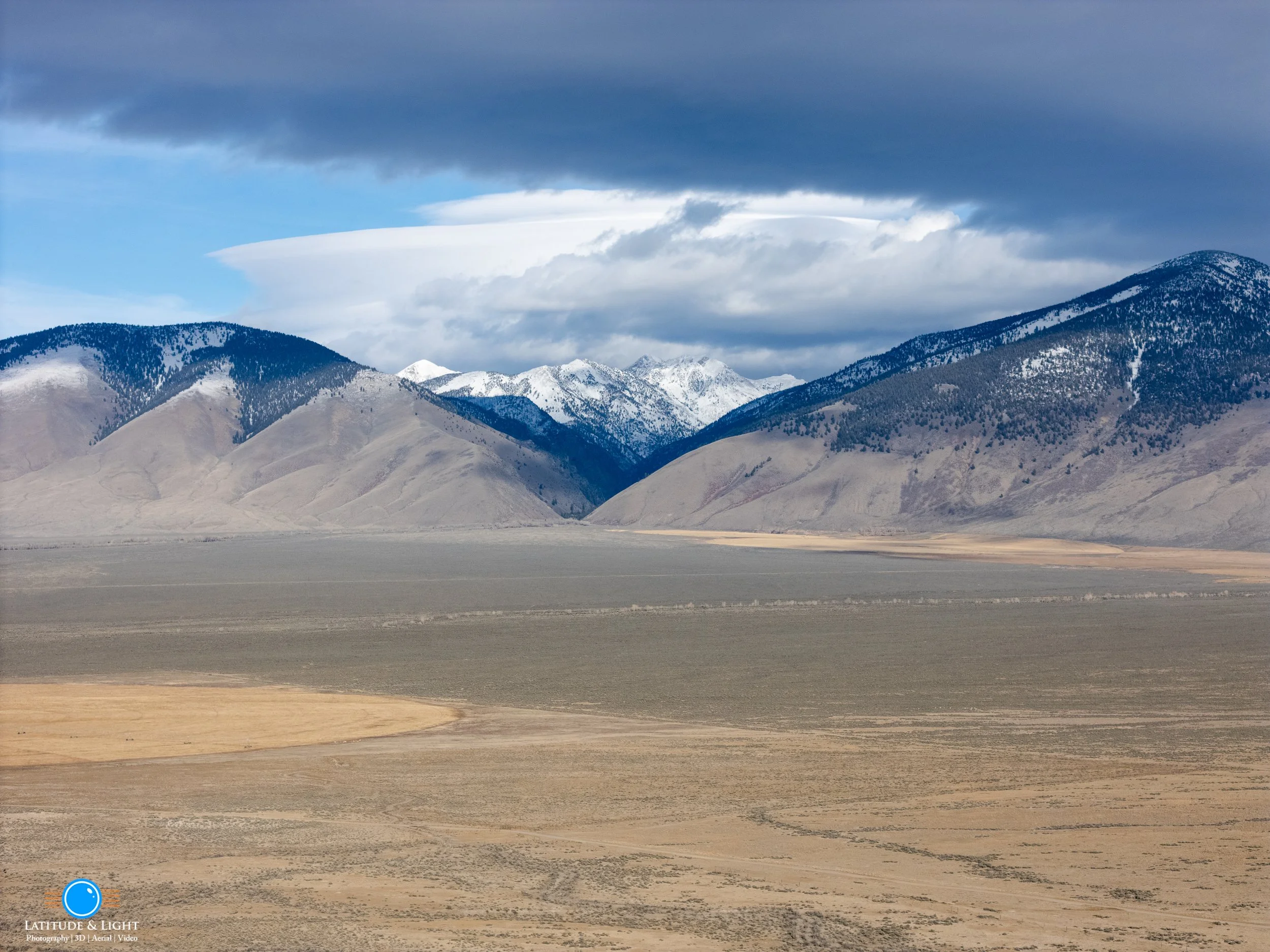 A wide landscape view of an Idaho  valley with the Lemhi mountains in the background, some of which are snow-capped, under a partly cloudy sky.
