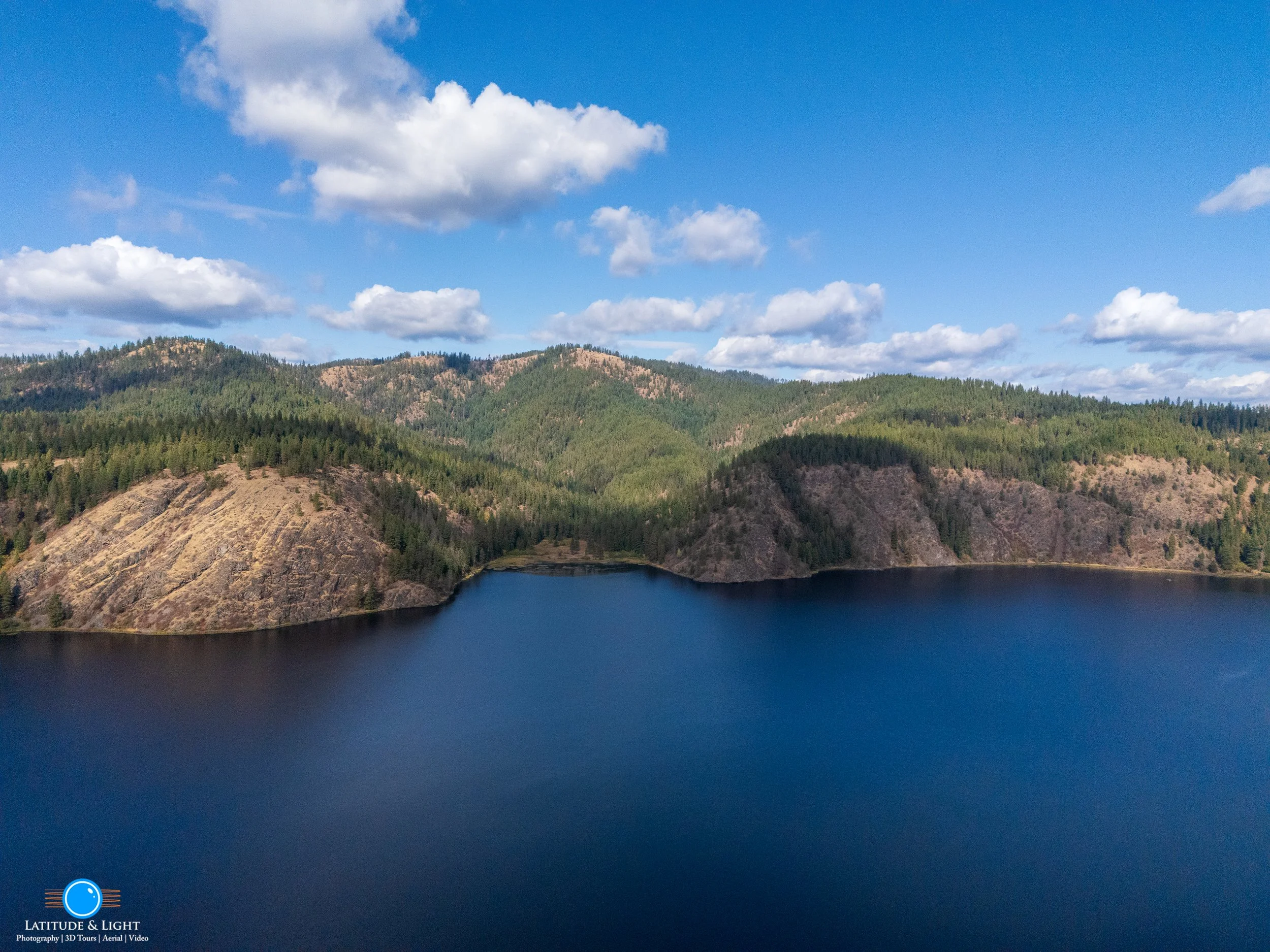 Harrison, Idaho: Aerial view of a lake surrounded by green forested hills and mountains under a partly cloudy sky.