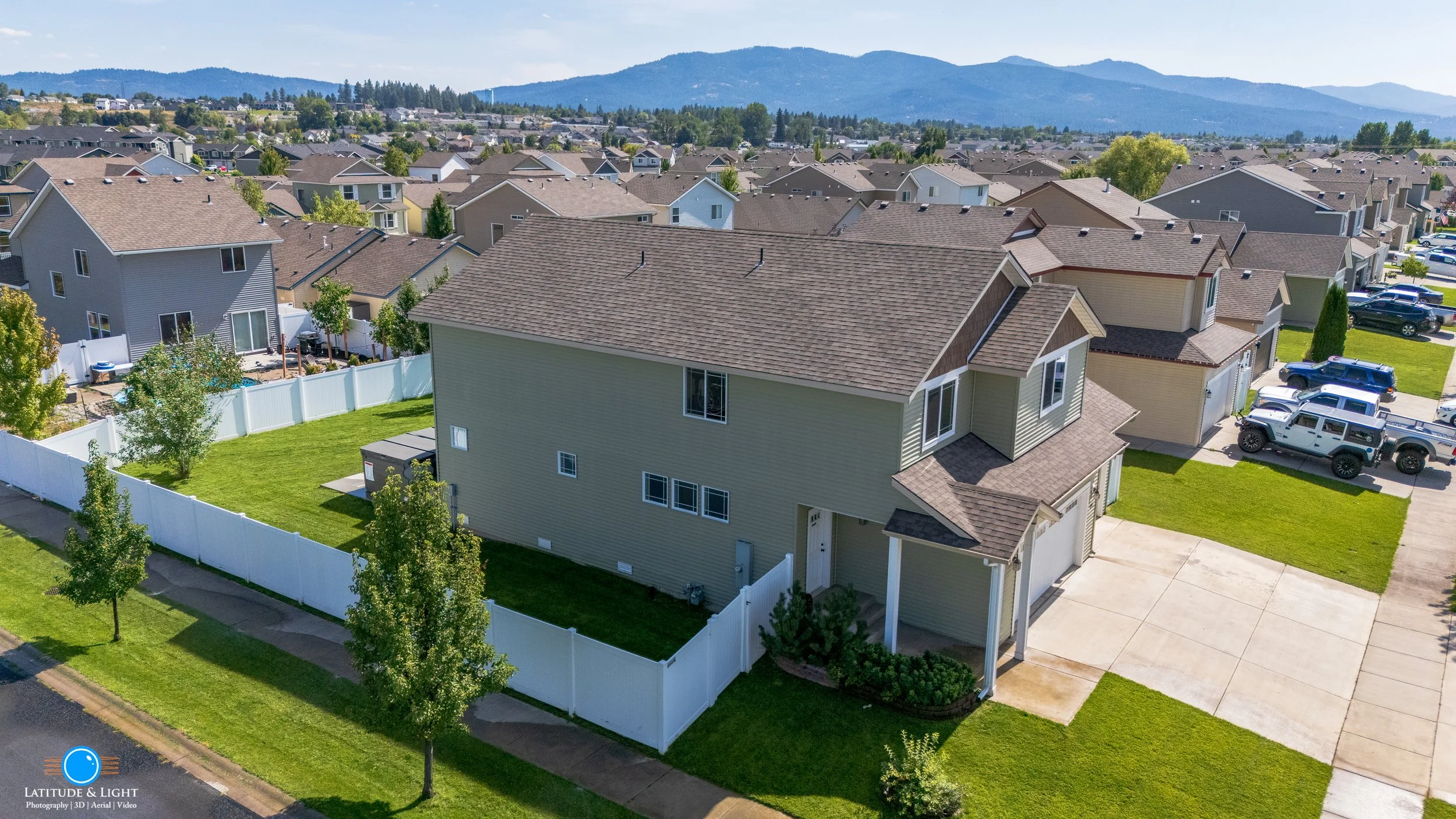 Aerial view of a suburban subdivision in Post Falls, Idaho with several houses, green lawns, and parked cars. Mountains are visible in the background.