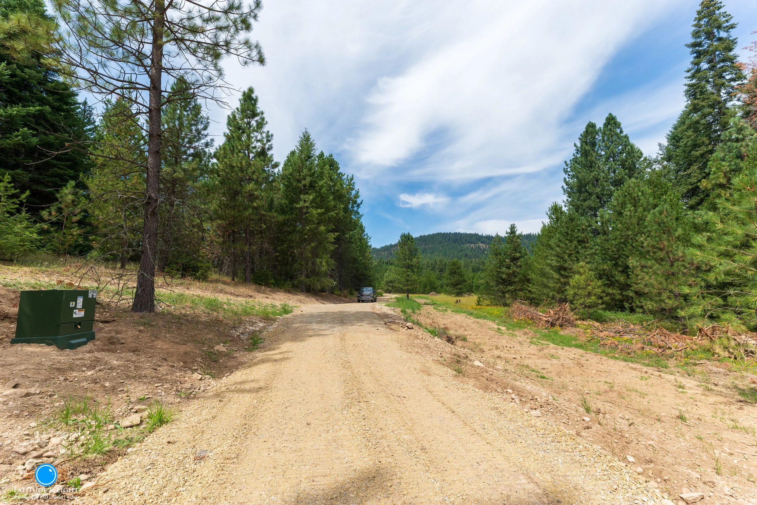Priest River, Idaho: A dirt road in a wooded area with pine trees on both sides, a black vehicle in the distance, and a blue sky with some clouds.