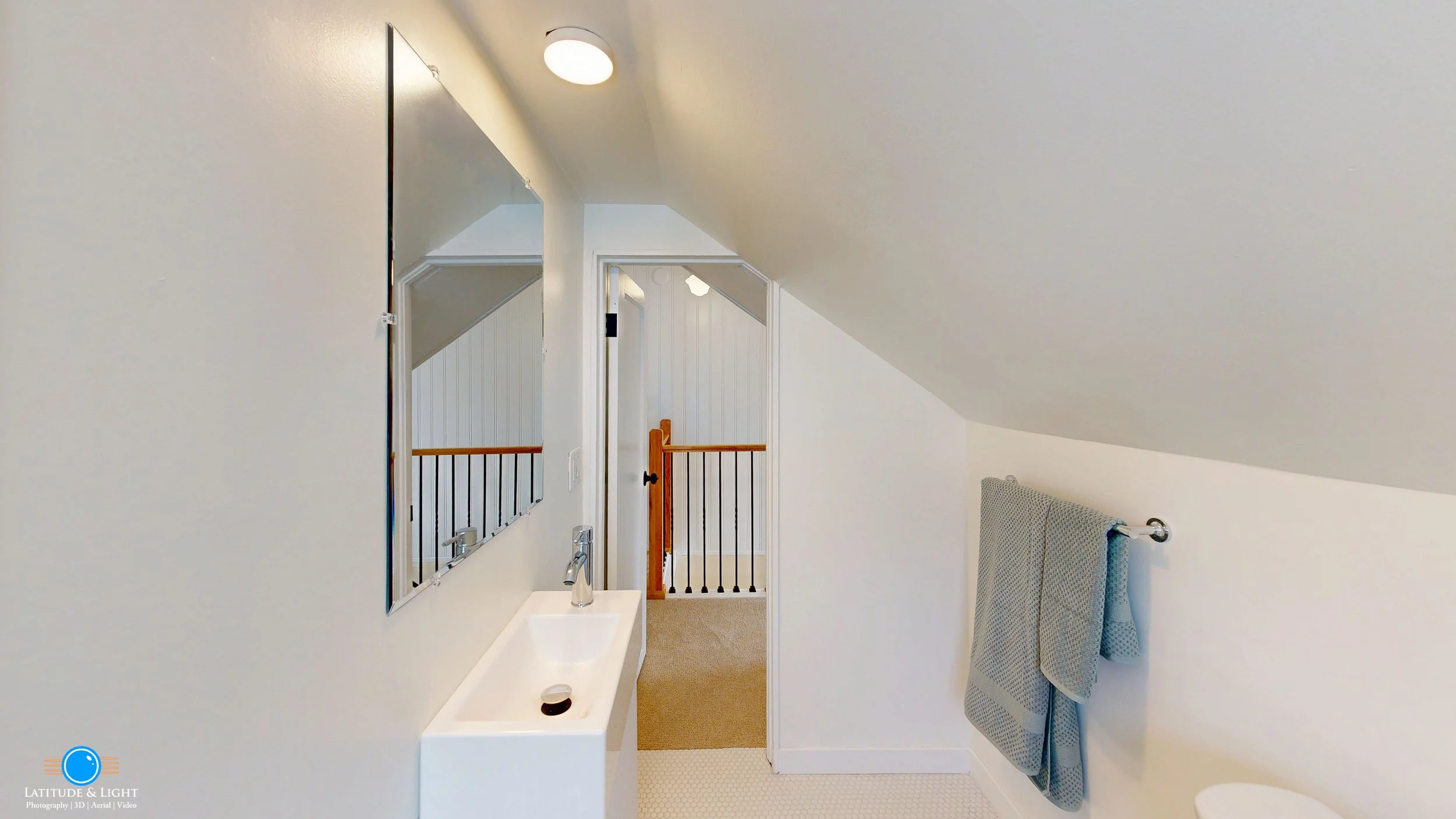 A small bathroom with a sloped ceiling, a rectangular mirror, a white sink with a chrome faucet, and two blue towels hanging on a towel bar. In the background, there is a door opening to a hallway with a wooden and black metal railing, white walls with beadboard, and a beige carpeted floor.