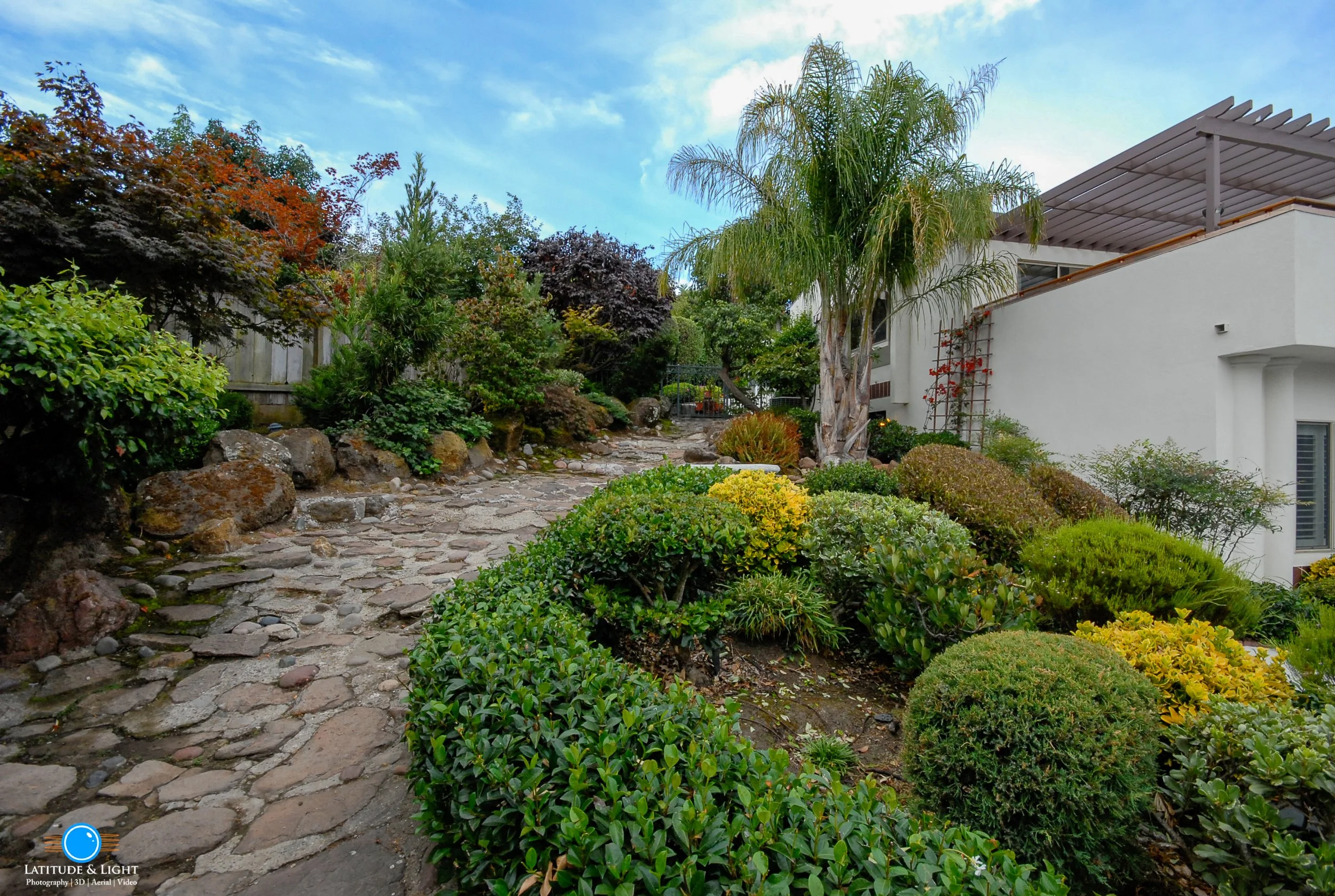 A stone pathway in a lush garden with various green shrubs, trees, and a Cupertino home with a balcony and trellis in the background.