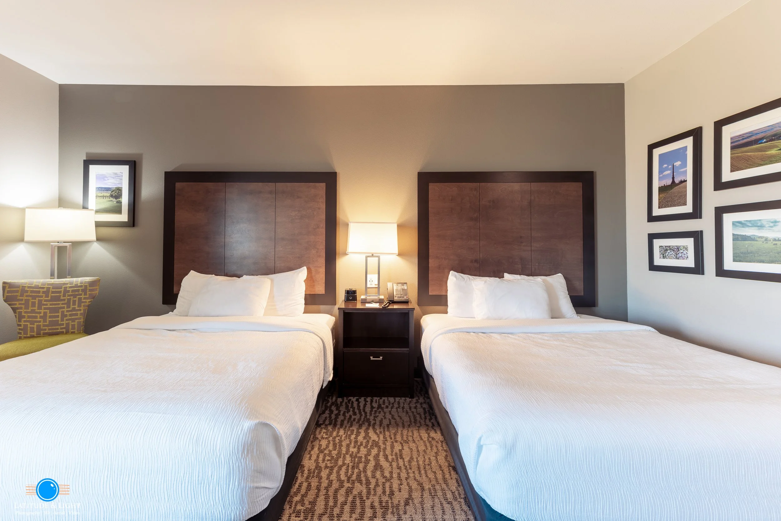 Hotel room with two beds, each with white bedding and pillows, centered nightstand with lamp, between the beds, gray painted walls, and framed pictures of landscapes on the wall.
