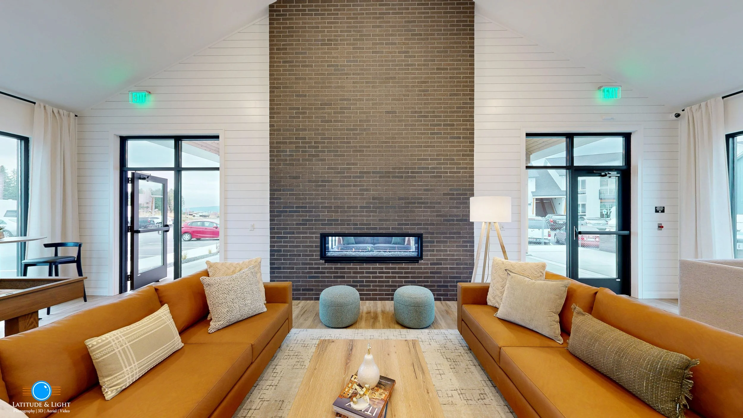 pokane Valley area apartment community center features two brown leather sofas, decorative pillows, a wooden coffee table, a central brick fireplace, and large windows with white curtains.