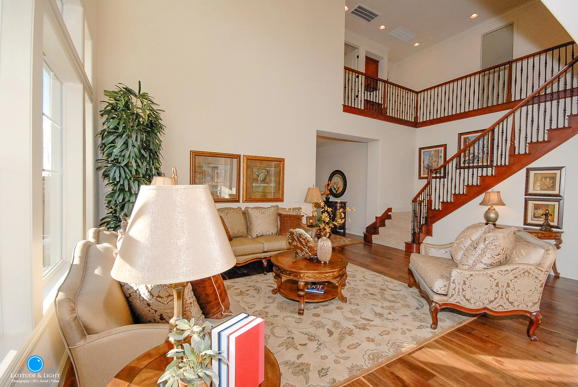 Living room with beige and cream furniture, wooden staircase, large window, lamps, framed artwork, and hardwood floors.