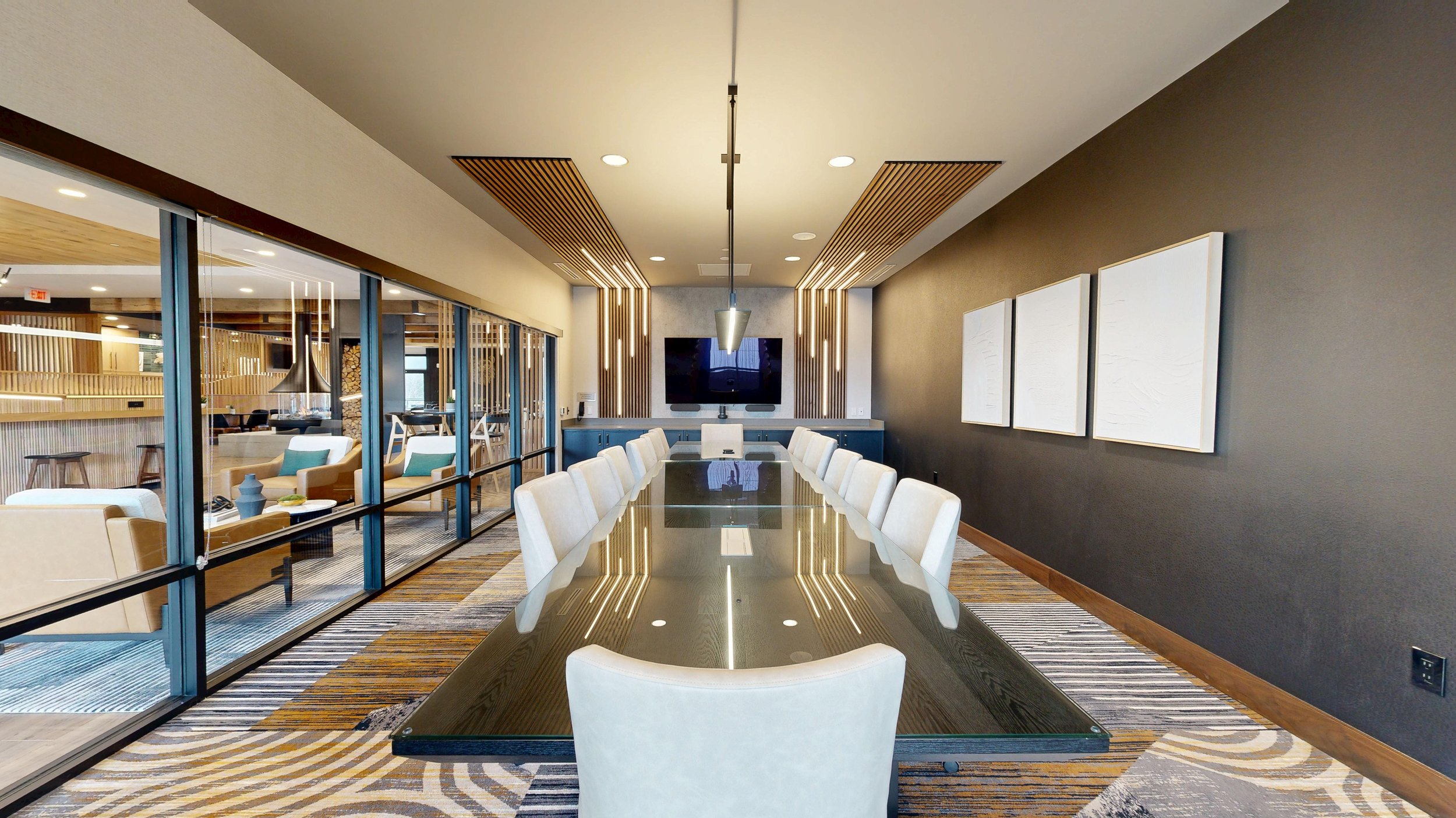 Modern hotel conference room in North Idaho with a long glass table surrounded by white chairs, large wall-mounted TV, abstract artwork, and a view into a lounge area with armchairs and stools.