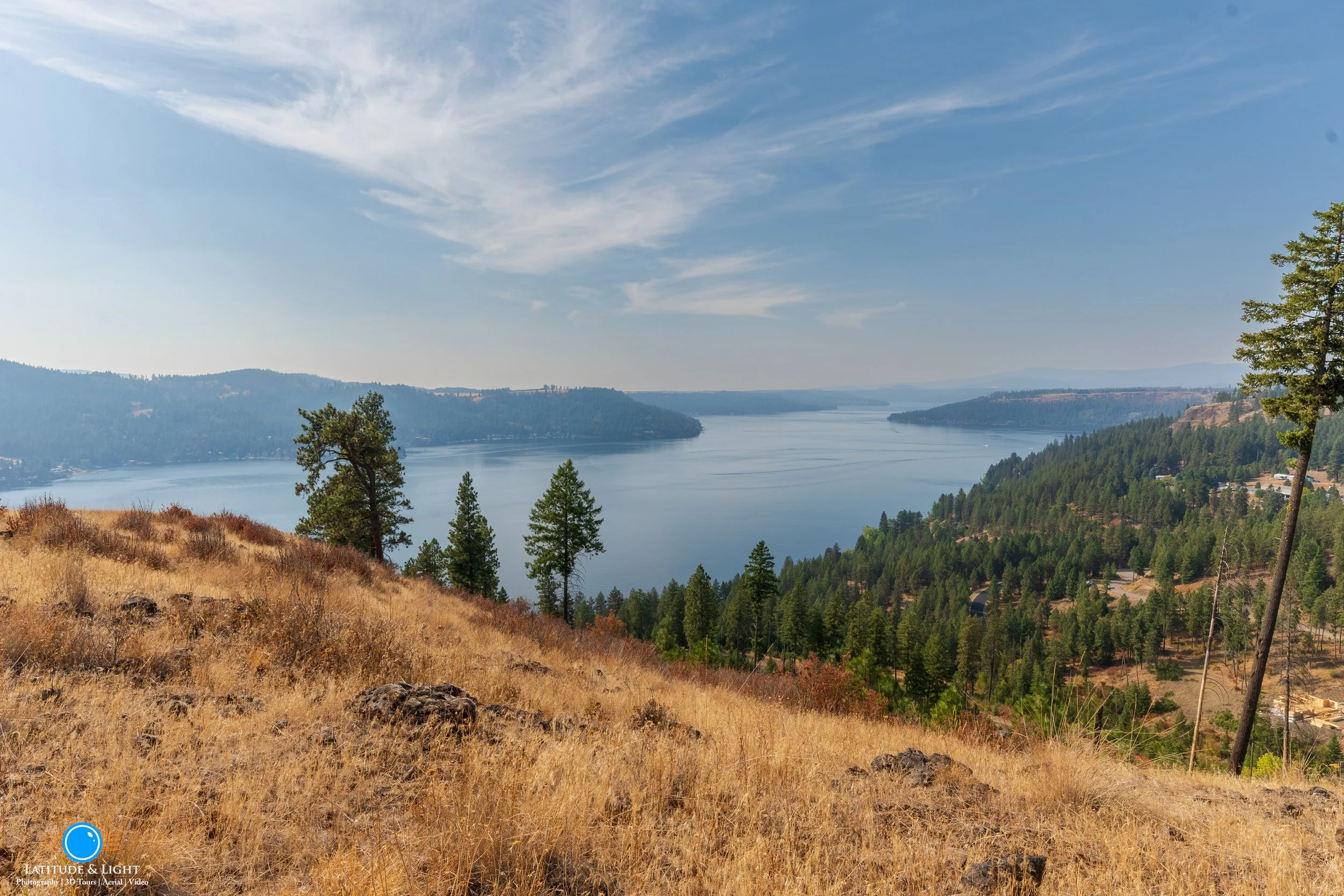Land in Harrison, Idaho: A scenic landscape with a large body of water, surrounded by forested hills and a dry grassy hillside in the foreground. The sky is blue with some clouds.