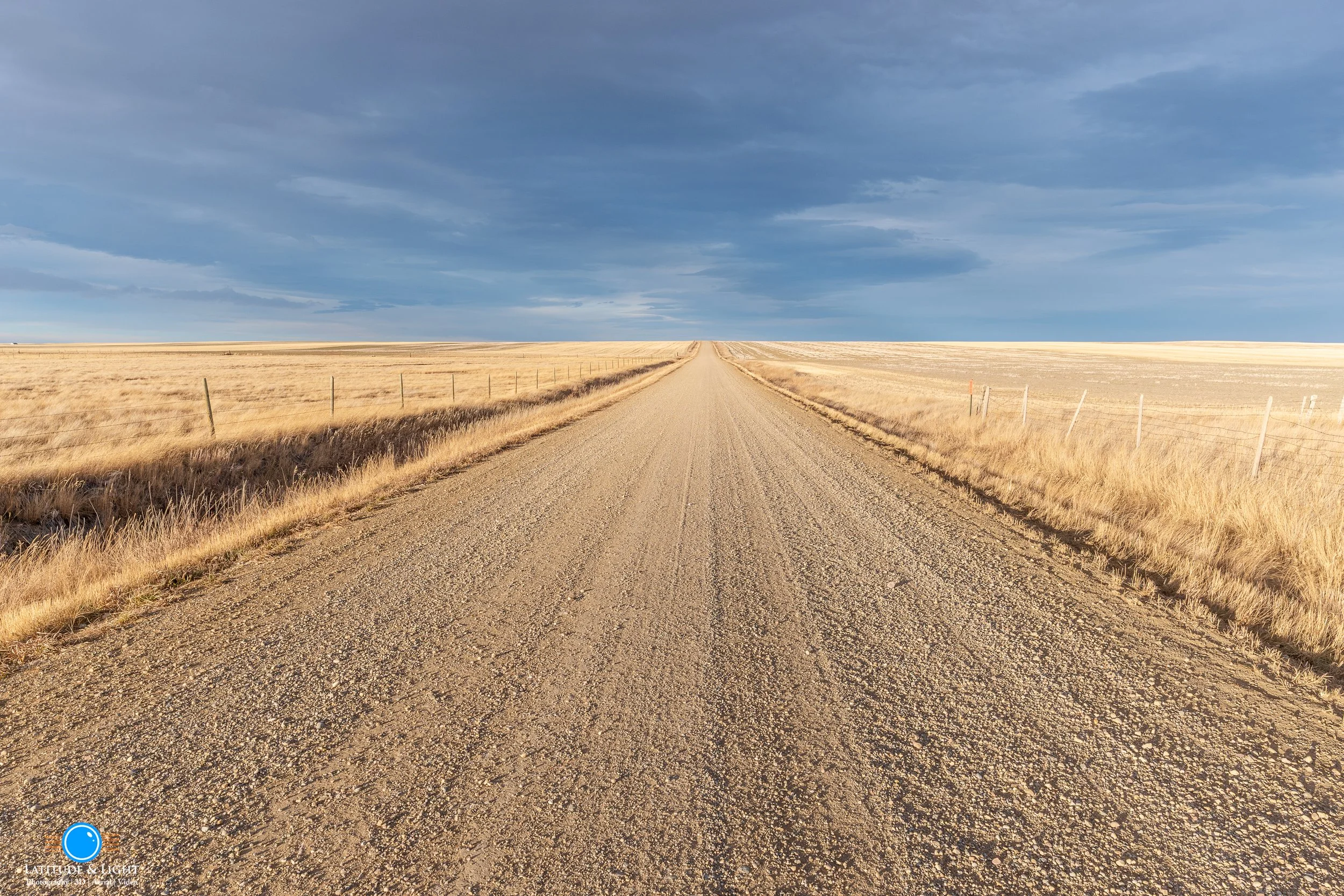 Dutton, Montana: A long dirt road stretching into the horizon through a flat, open field with dry, yellow grass. The sky is partly cloudy with a mix of dark and light clouds.