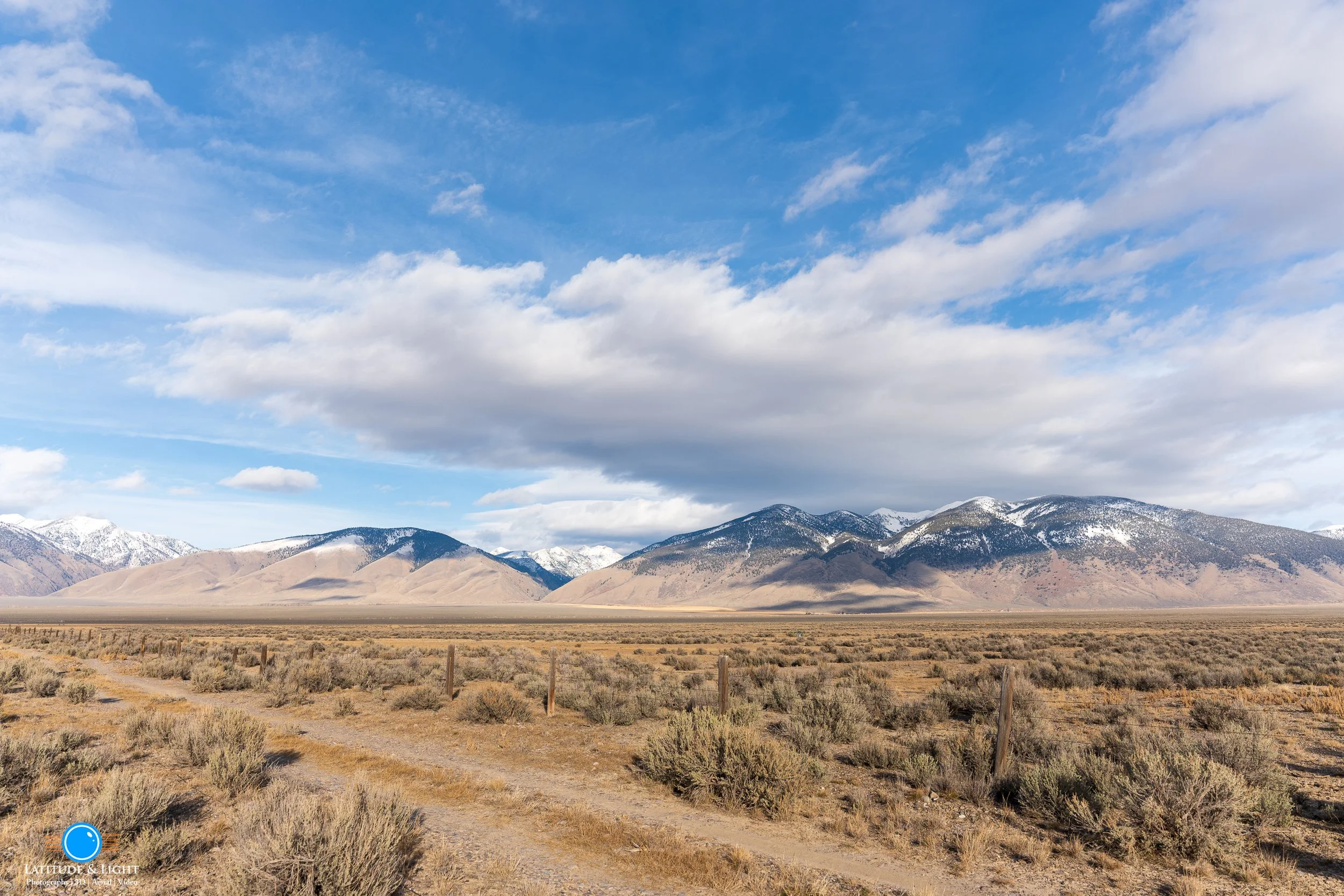 Open desert landscape with dirt path, sparse shrubbery, and Idaho's Lemhi mountain range with snow-covered peaks under a sky with scattered clouds.