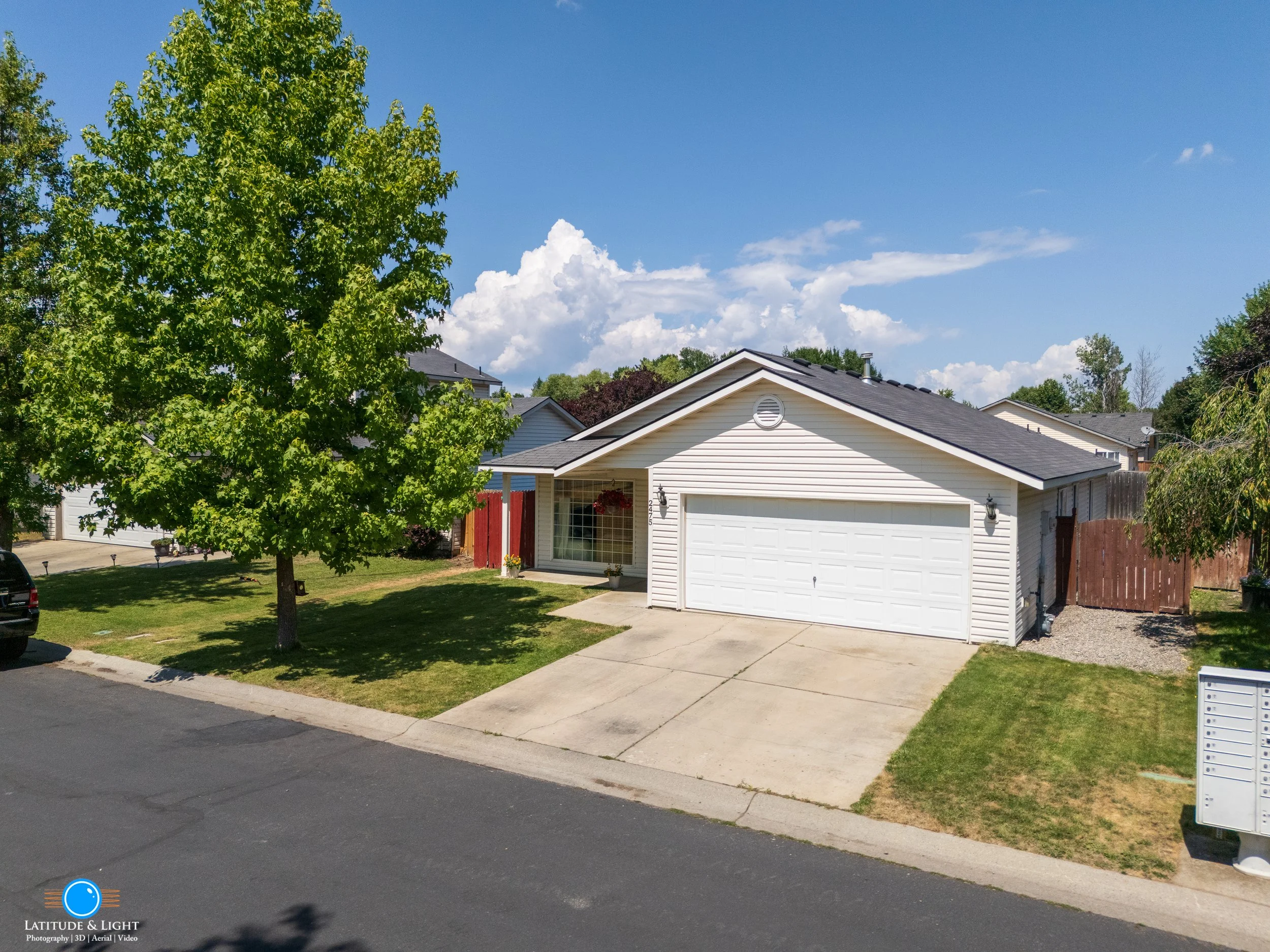 A suburban Coeur D'Alene house with a white garage door and a large green tree in the front yard, under a partly cloudy sky.