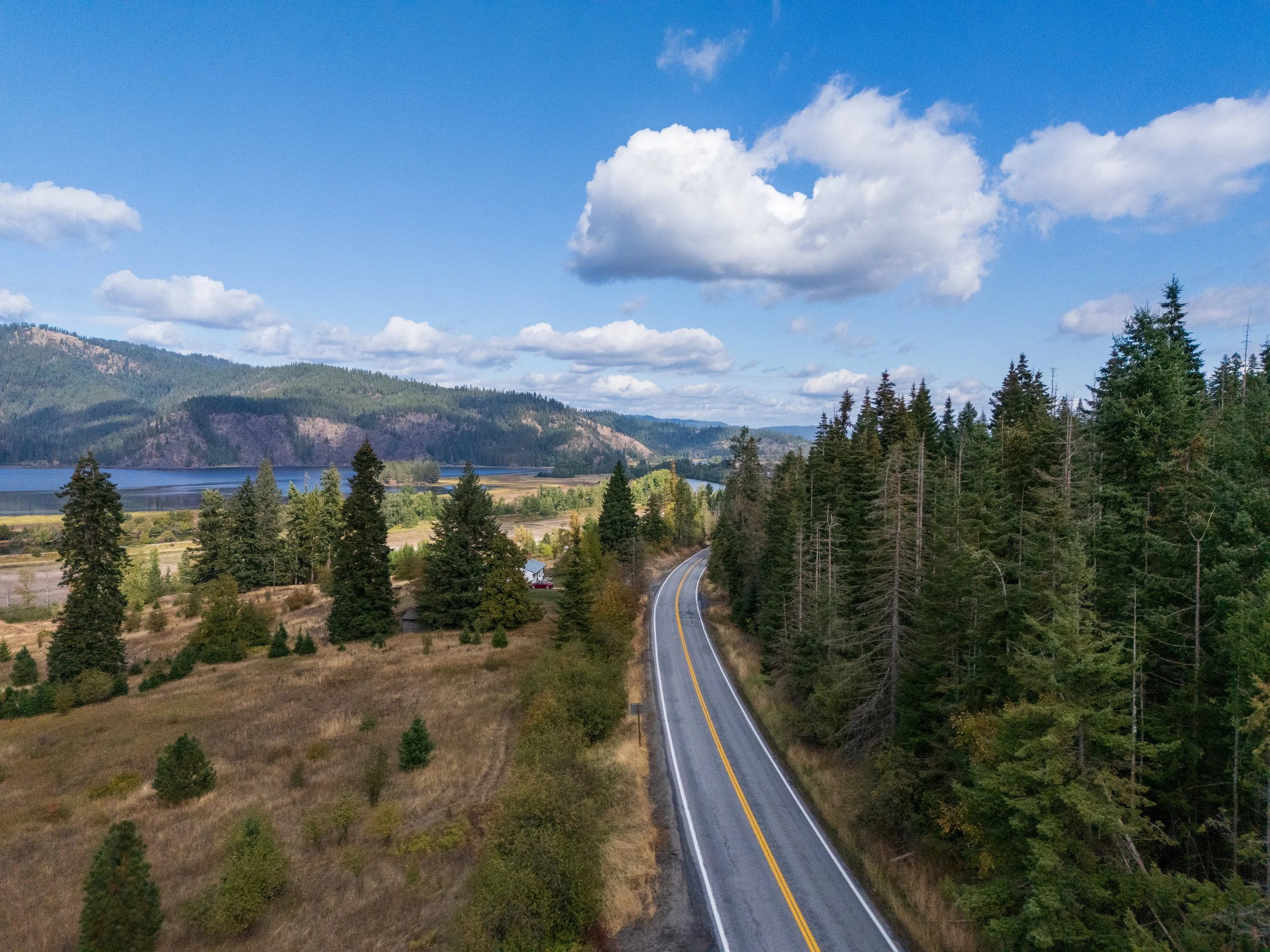 A winding road runs through a forested area in Harrison, Idaho near a body of water with mountains in the distance under a partly cloudy sky.