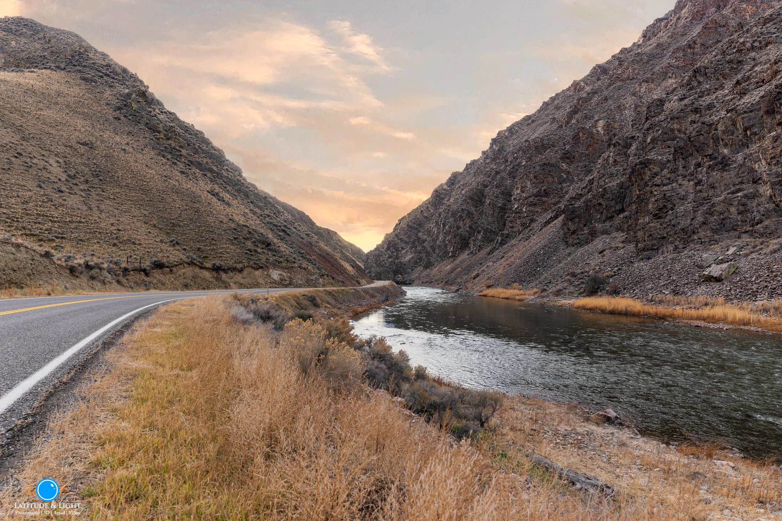 A scenic view of a winding Idaho Highway 93 running alongside the Salmon River with steep rocky hills on both sides under a partly cloudy sky at sunset or sunrise.