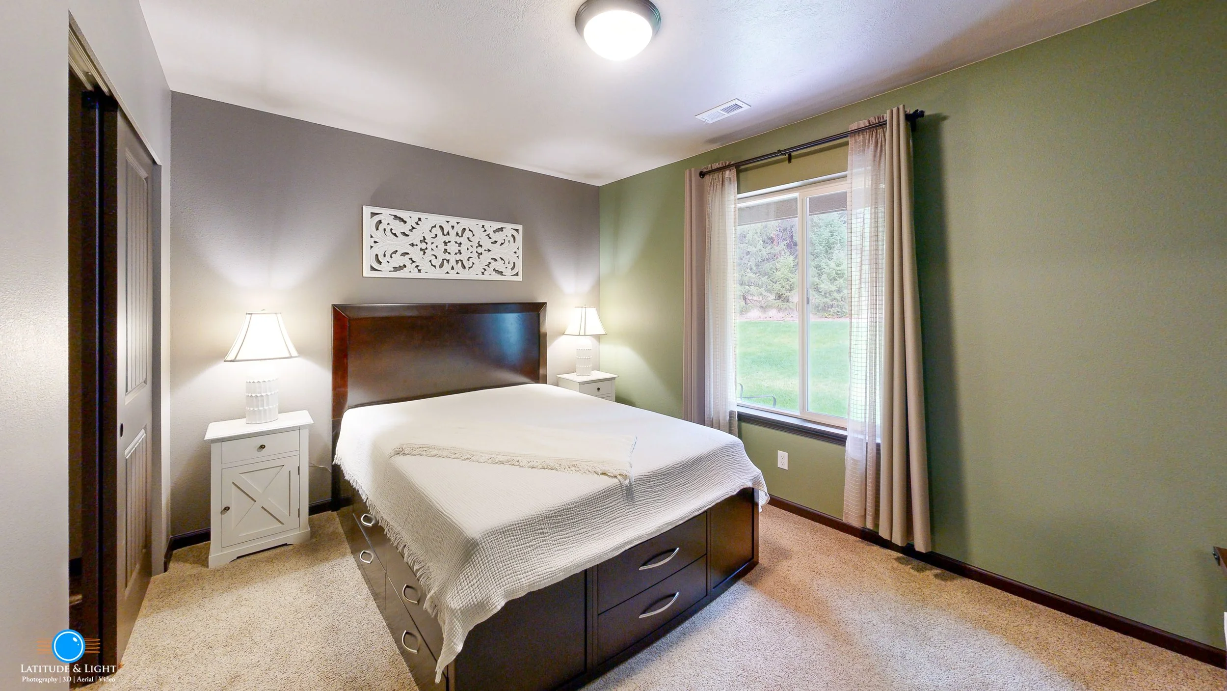 North Idaho bedroom with a large bed, two white nightstands with lamps, a window with beige curtains, and a gray and green painted wall.