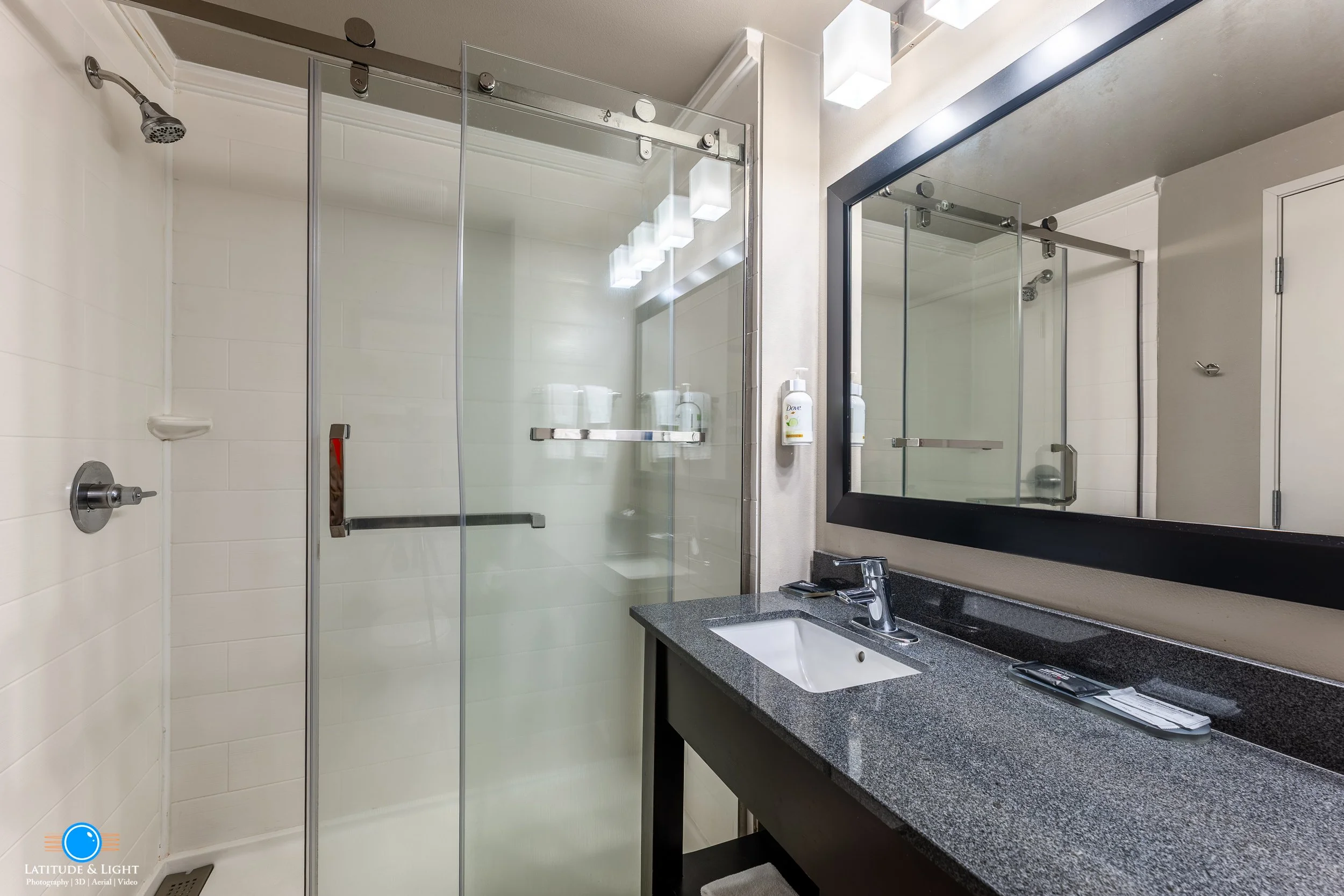 Bathroom with a glass shower enclosure, a black-framed mirror, a gray countertop with a sink, and a soap dispenser.