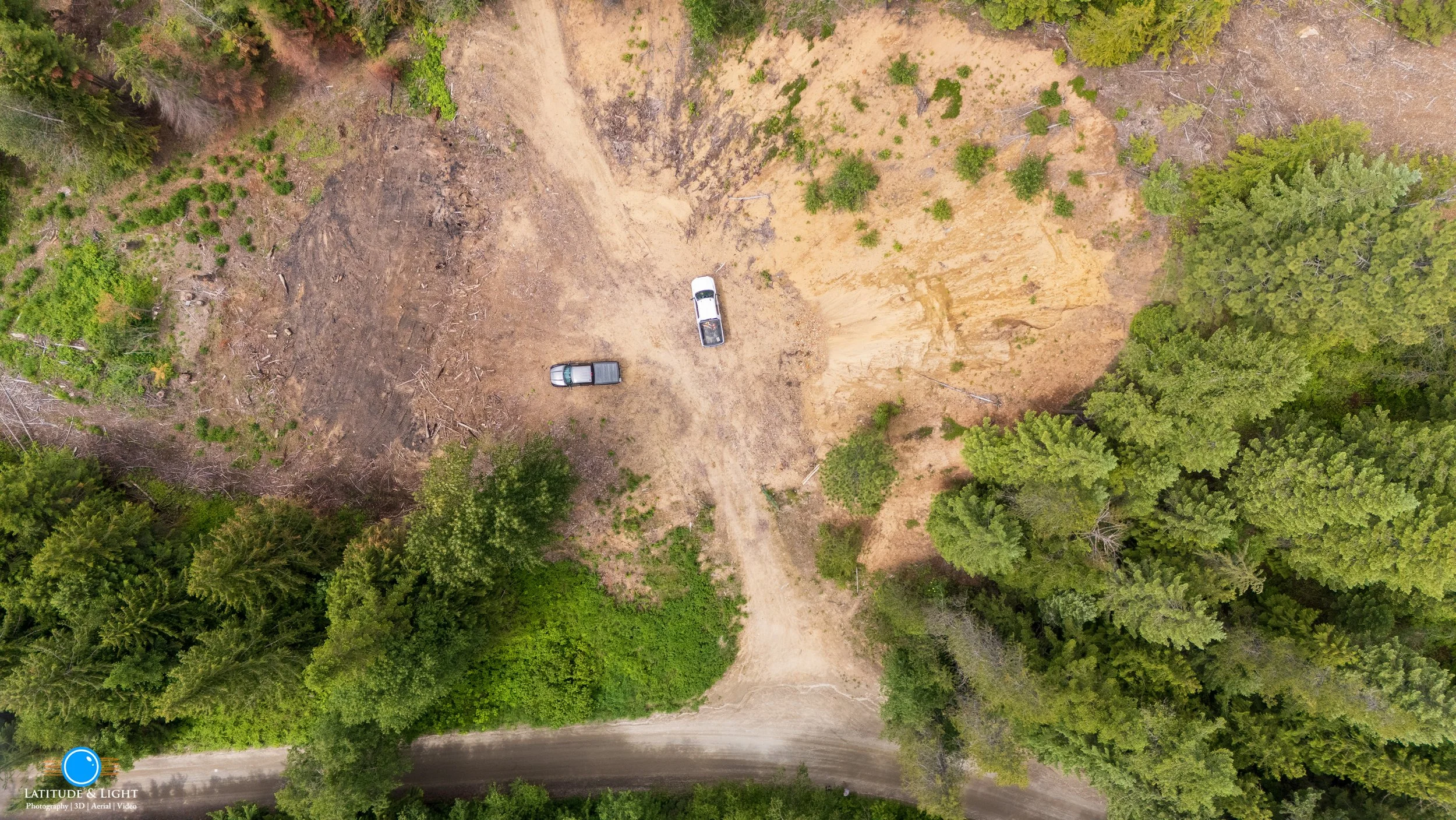 North Idaho land: An aerial view of a cleared patch of land with two parked vehicles, surrounded by dense green forest, and a dirt road at the bottom.
