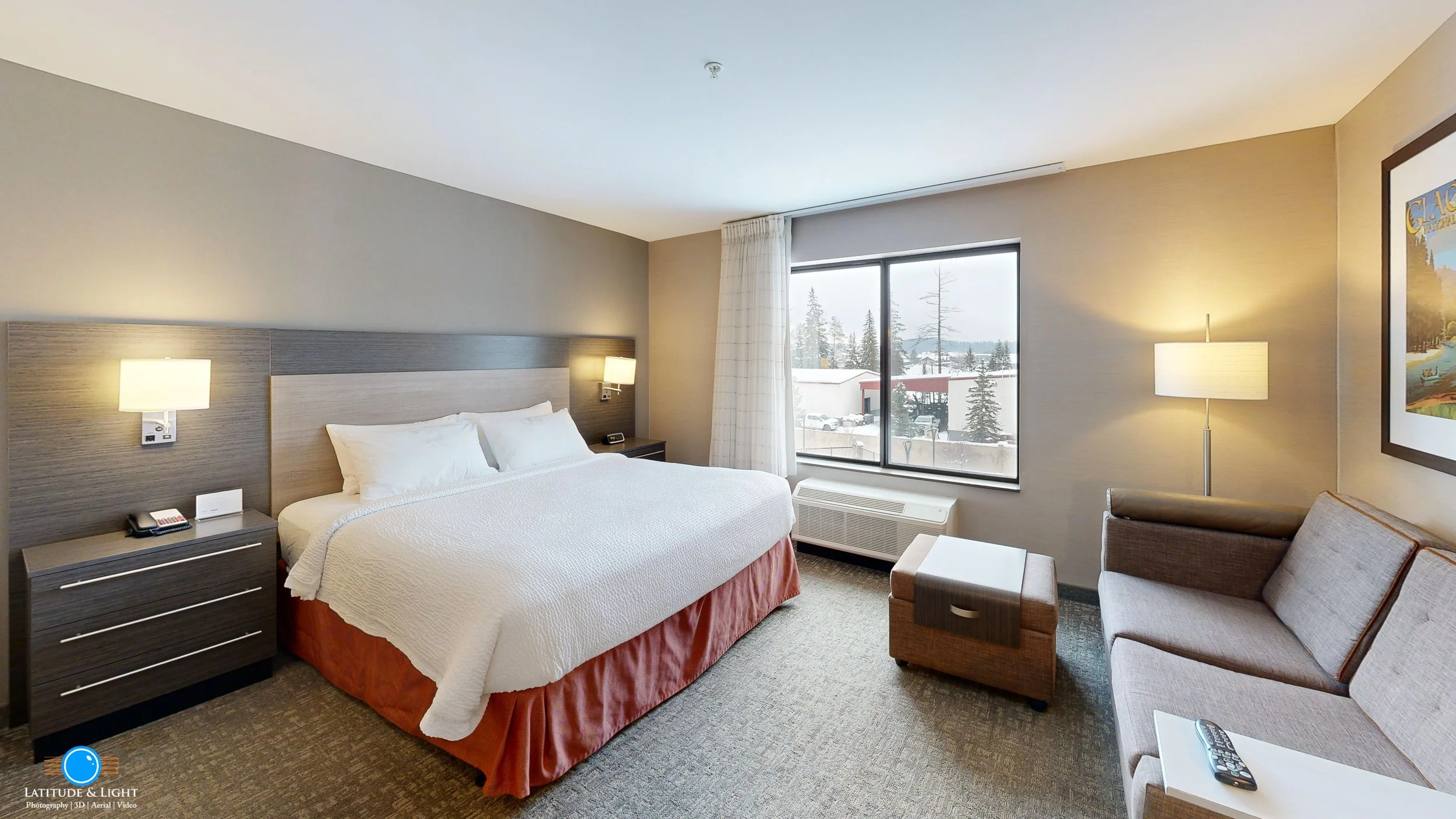 A hotel room with a large bed, nightstand, sofa, table, window with curtains, and outside views of snow and trees.